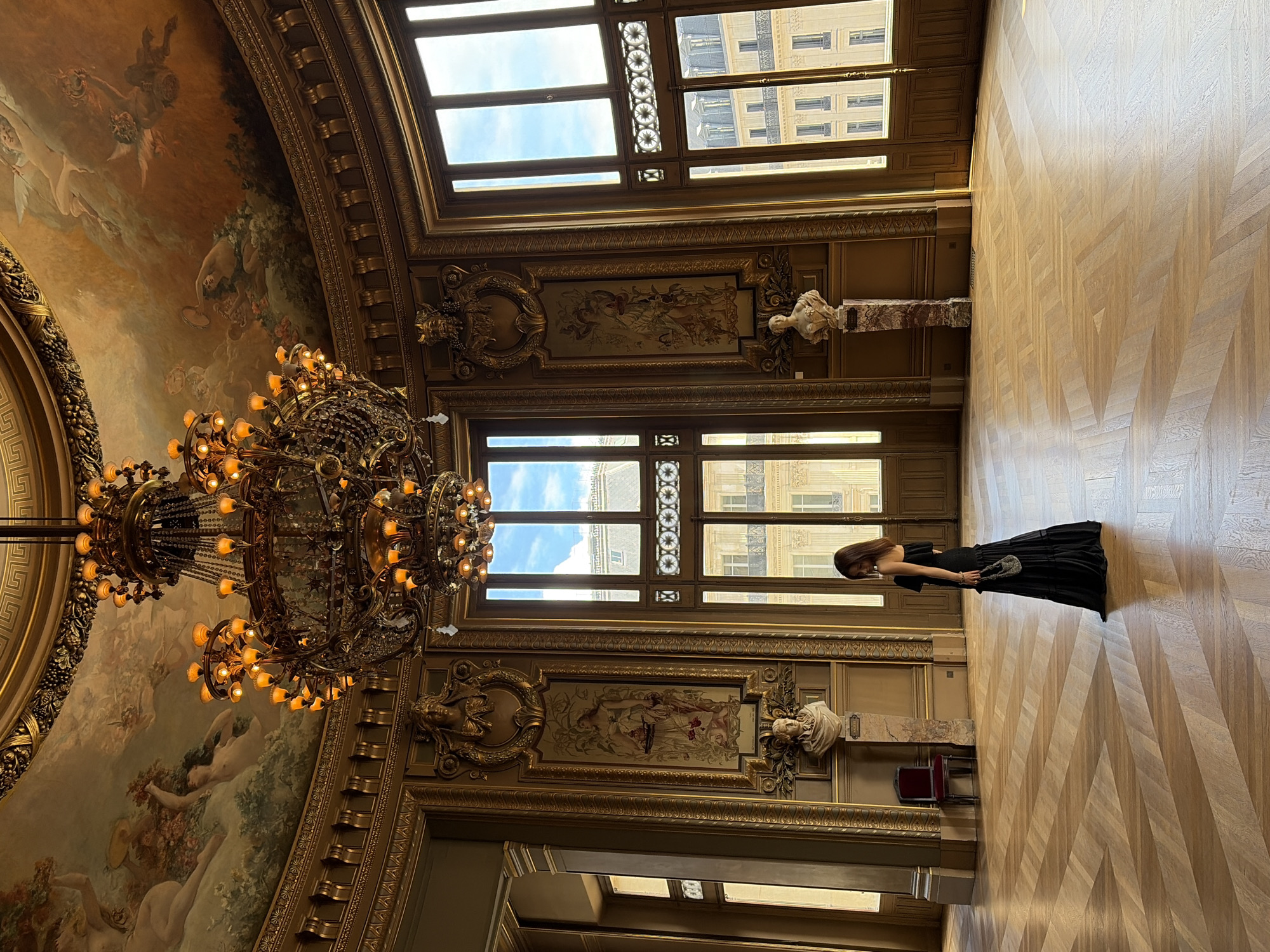 In a grand, ornate hall, likely within a Parisian opera house or museum, a lone woman stands on a polished wooden floor. She is dressed in a striking, floor-length black gown and clutches a small, beaded purse. The room is richly decorated with gilded accents, intricate moldings, and large, classical paintings on the ceiling and walls, depicting mythological scenes with cherubic figures. A massive, multi-tiered crystal chandelier hangs from the ceiling, its numerous lights casting a warm glow. Large windows, divided into multiple panes, offer views of a bright, daytime sky and the architecture of surrounding buildings. The floor is laid with light-colored wooden planks in a striking herringbone pattern, reflecting the light. Busts of classical figures are mounted on the walls. The scene suggests a moment of quiet contemplation or admiration within a historically significant and opulent space.
