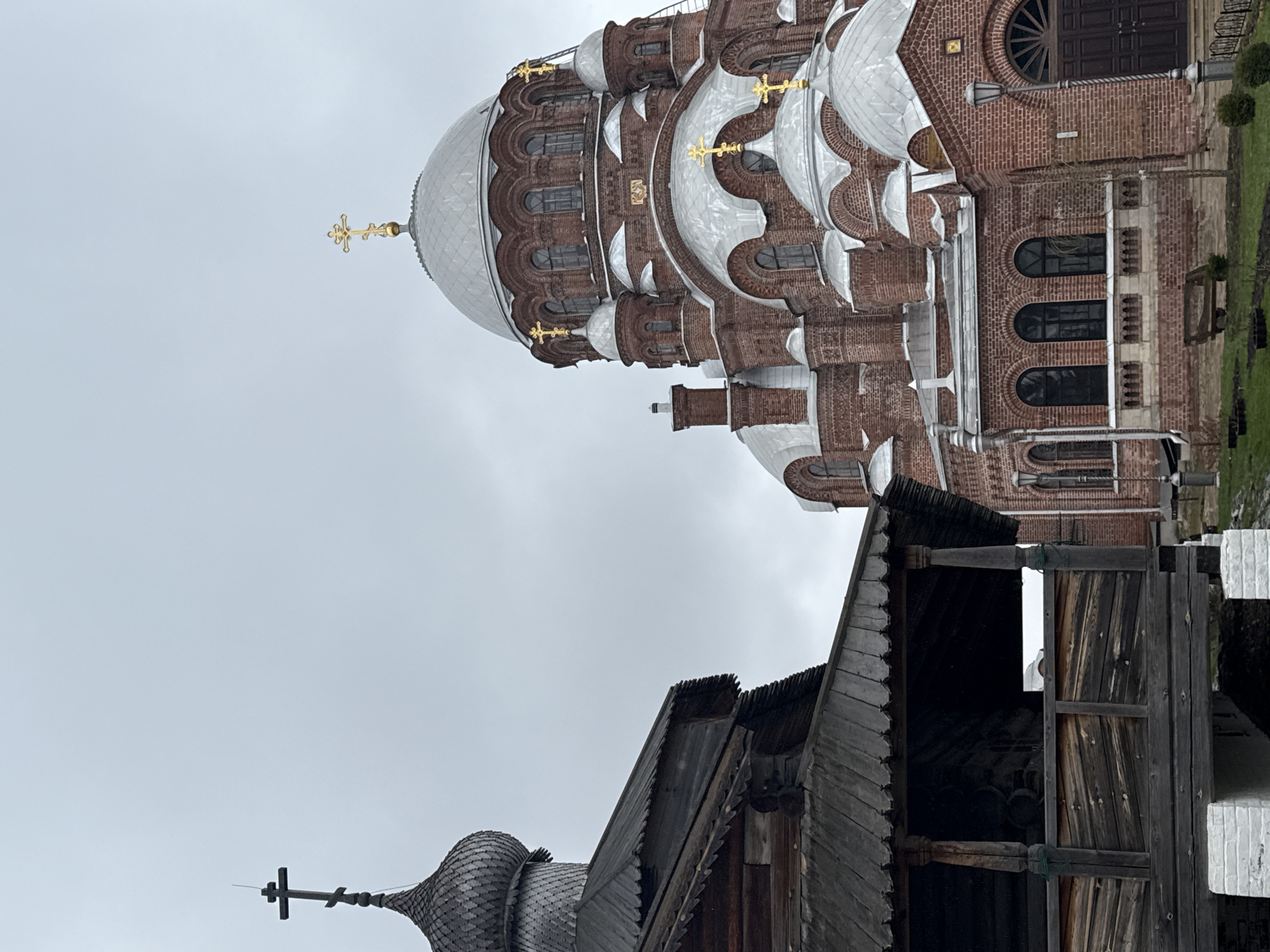 The image captures a view of two Orthodox churches in Sviyazhsk, Russia, under an overcast sky. In the foreground, a dark wooden structure with a layered roof and a cross at the apex is visible. Behind it, a larger, brick-built cathedral with silver domes and gold crosses stands prominently. The cathedral features detailed architectural elements such as arched windows and decorative brickwork. Some greenery can be seen on the right side of the image. The sky is uniformly gray, suggesting a cloudy day.
