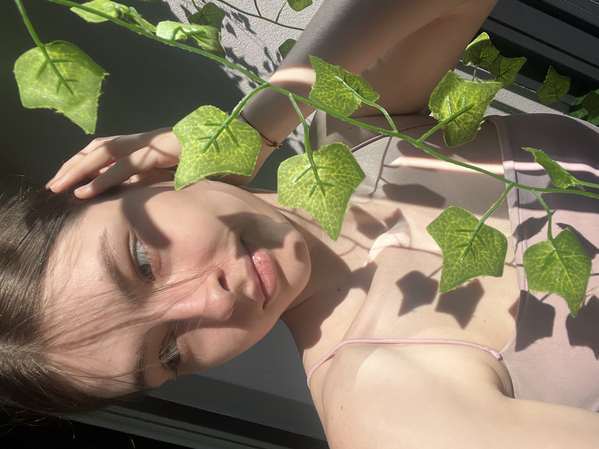 This media file features a close-up, overhead shot of a young woman lying down, her face tilted upwards. She has fair skin, light brown hair, and appears to be wearing a light pink camisole. Artificial green ivy vines with leaves are draped across her and the scene. Sunlight casts dappled shadows on her skin and the wall behind her, indicating a bright, sunny day. The overall mood is serene and relaxed.
The woman's hand rests on her head, and her eyes are partially closed, suggesting a moment of calm or repose. Her lips are slightly parted, and she has a soft expression. The lighting suggests it is daytime, likely late morning or early afternoon, given the angle of the shadows. The background appears to be an outdoor setting, possibly a balcony or patio, with a textured wall and a hint of dark horizontal blinds in the upper right corner. There is no visible text in the image. The context of Sinyavino, Russia, provides a geographical location but does not add specific visual details to the scene itself.