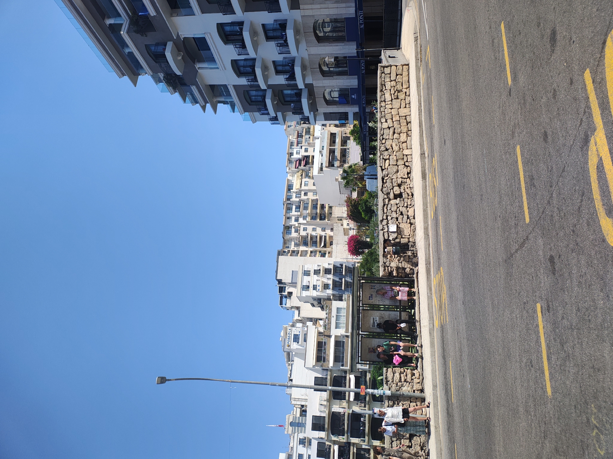 A sunlit, eye-level medium shot shows a street scene in Mellieħa, Malta. The foreground is dominated by a paved road with yellow road markings. Behind the road is a bus stop with a stone wall and shelter. Five people are visible at the bus stop, some sitting and some standing. Two individuals stand outside the shelter, facing away from the camera. Behind the bus stop, a cluster of white and beige apartment buildings rises against a clear blue sky. To the right, a modern, multi-story building with balconies and glass railings stands prominently. The overall impression is one of a typical urban environment on a bright, clear day.