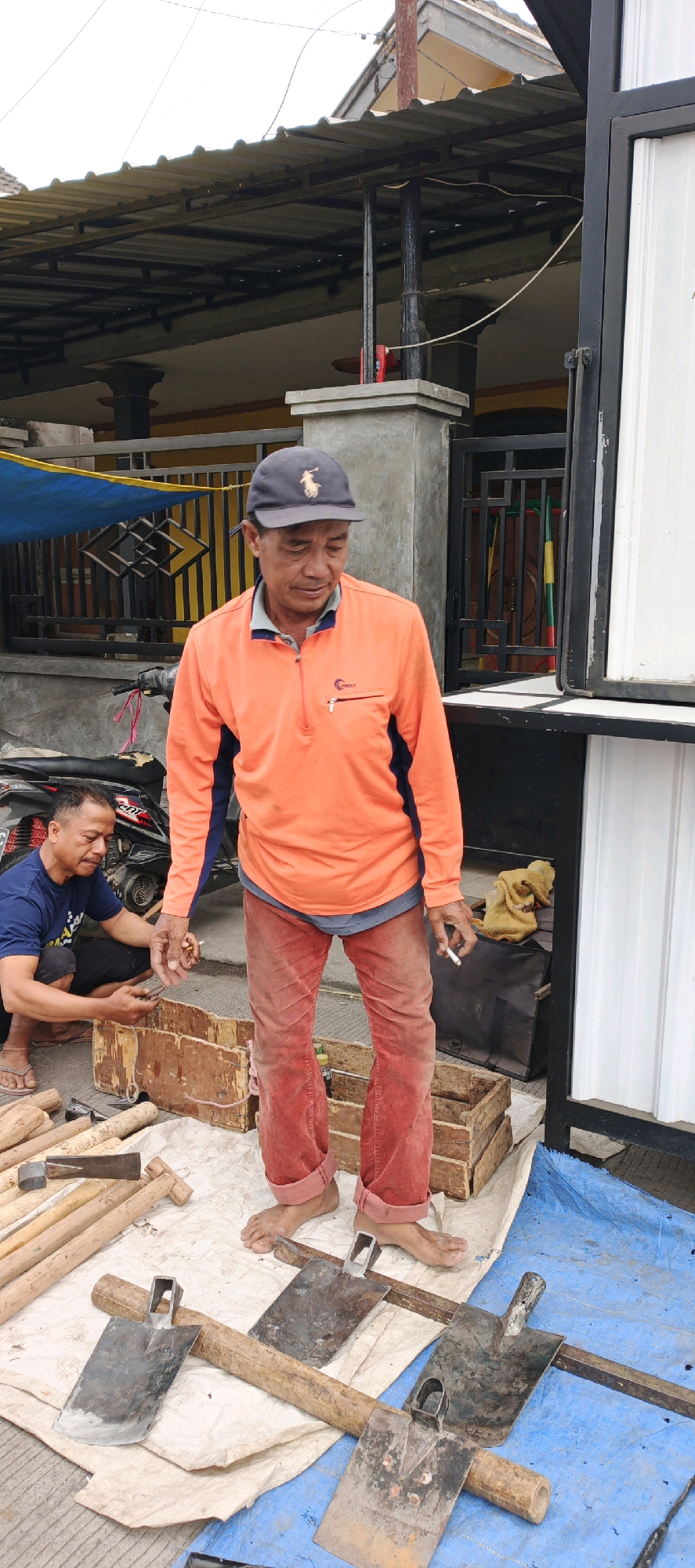 In Kasiyan, Indonesia, two men are gathered outdoors. The man in the foreground, wearing an orange long-sleeved shirt and red pants, stands facing slightly away from the camera with a cigarette in his right hand. In front of him, a collection of agricultural tools, including hoes and axes, are laid out on a white tarp. The tools have wooden handles and metal heads, some appearing worn and rusted. Wooden crates are stacked behind the tools. To the left, a second man, wearing a blue t-shirt and dark shorts, is crouched down and appears to be examining something in his hands. He is also holding a cigarette. The background shows a building with a corrugated metal roof and a fence. A motorcycle is parked to the left of the standing man. The ground is paved, and a blue tarp is visible in the lower right corner of the image. The scene appears to be an outdoor market or roadside stall selling farming equipment.