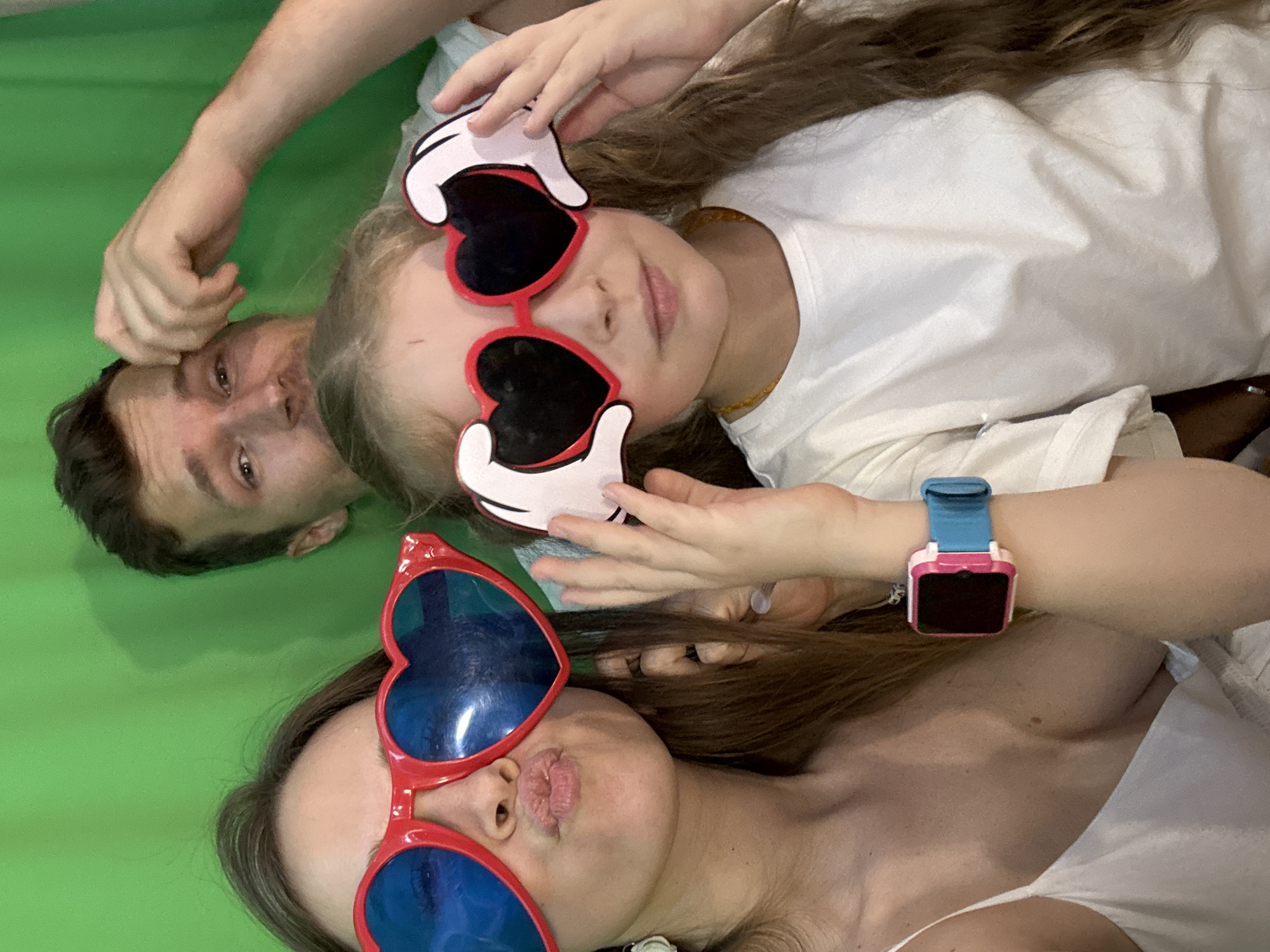 The image shows three people lying on a green surface. A man's head is visible in the upper left corner. A young girl wearing heart-shaped sunglasses with Mickey Mouse glove designs is centrally positioned, looking at the camera. A woman wearing red heart-shaped sunglasses with blue lenses is positioned below the girl, also looking at the camera and puckering her lips. The woman is wearing a blue and pink watch. The location is not discernible from the image.