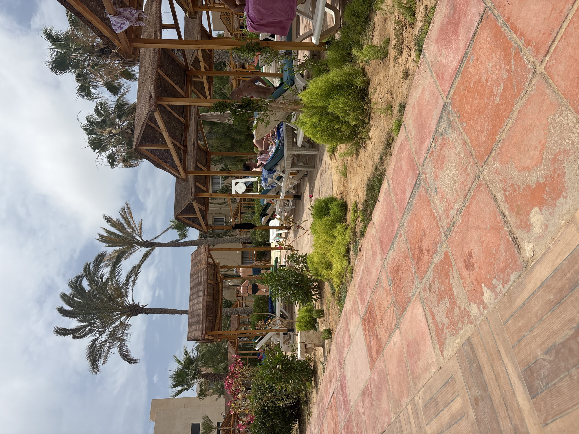 In Taguermess, Tunisia, a resort area features a tiled walkway in the foreground leading to a sandy area with loungers under thatched-roof cabanas. Palm trees and green foliage are visible. Several people are relaxing on loungers, some partially obscured by greenery or the cabana structures. One couple stands near a cabana, while others are seated or lying down. The sky is cloudy.