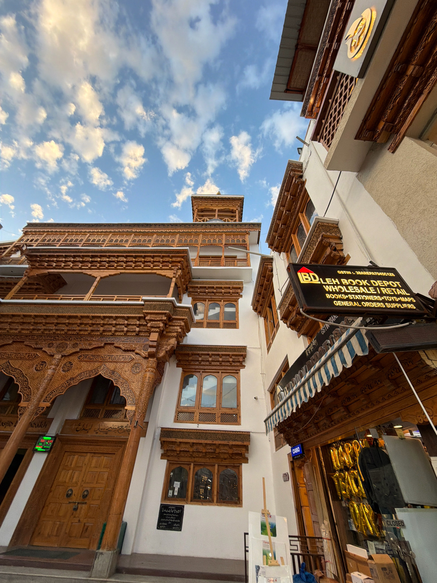 The image shows the Leh Book Depot, a wholesale and retail establishment in Leh, India. The store sells books, stationery, toys, maps, and general orders. The facade features intricate wooden carvings and balconies, typical of Ladakhi architecture. A sign with the store's name and a list of its offerings is prominently displayed. In the foreground, an awning shades the entrance, and items like stacked boxes and a "CLOSED" sign are visible. The background is dominated by a clear blue sky with scattered white clouds. The shot is taken from a low angle, looking upwards at the building.