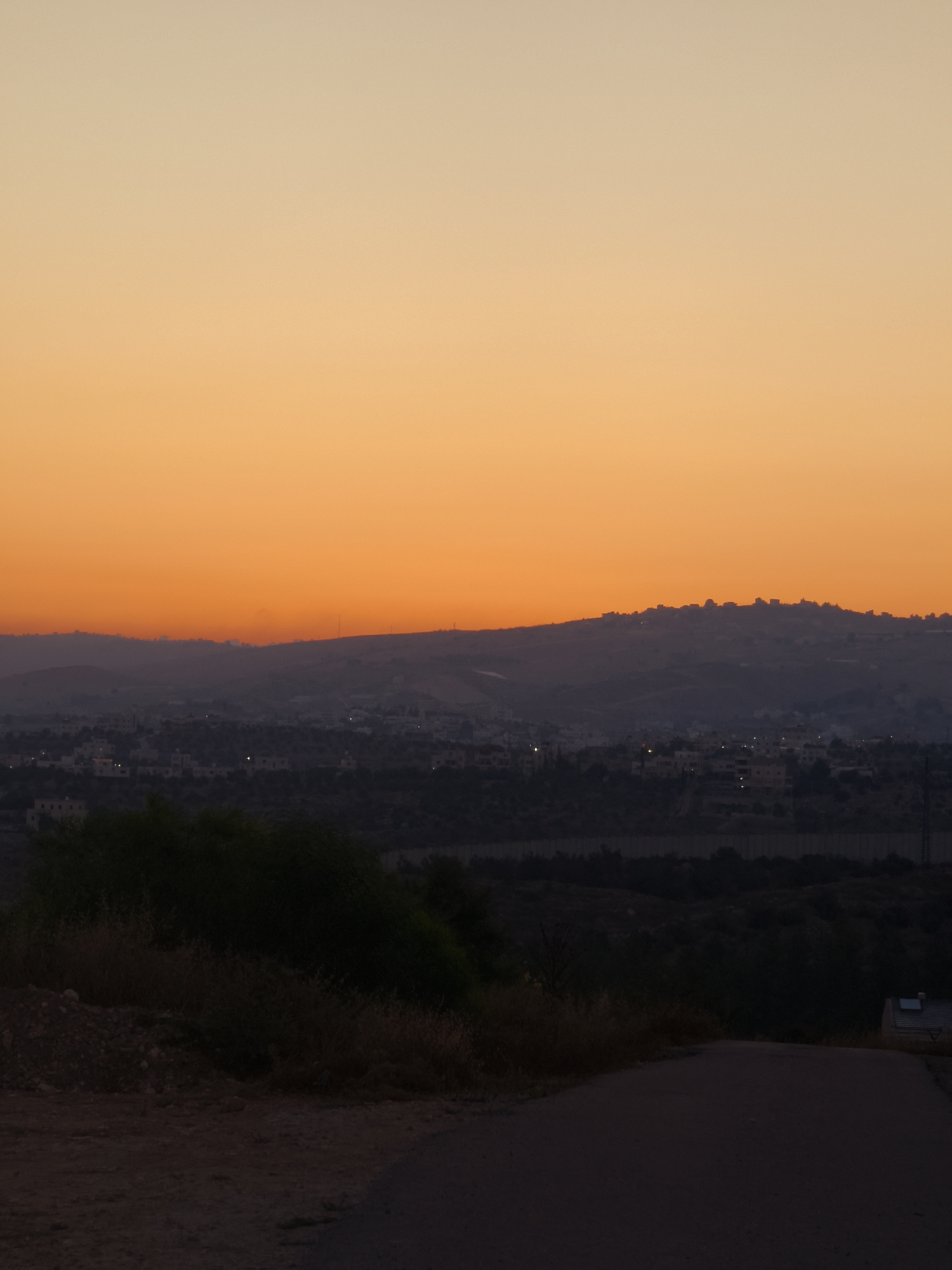 A wide shot captures a landscape at dusk, featuring a gradient sky of orange and yellow. Below the sky, a silhouette of hills is visible, with a town nestled in the valley. The town has scattered lights indicating habitation. In the foreground, a dark, unpaved road curves upwards. To the left of the road, a dense cluster of dark green bushes and dry grass is present. The image appears to be taken from an elevated position, looking out over the town and hills. This landscape is located within the Lachish Regional Council in Israel.