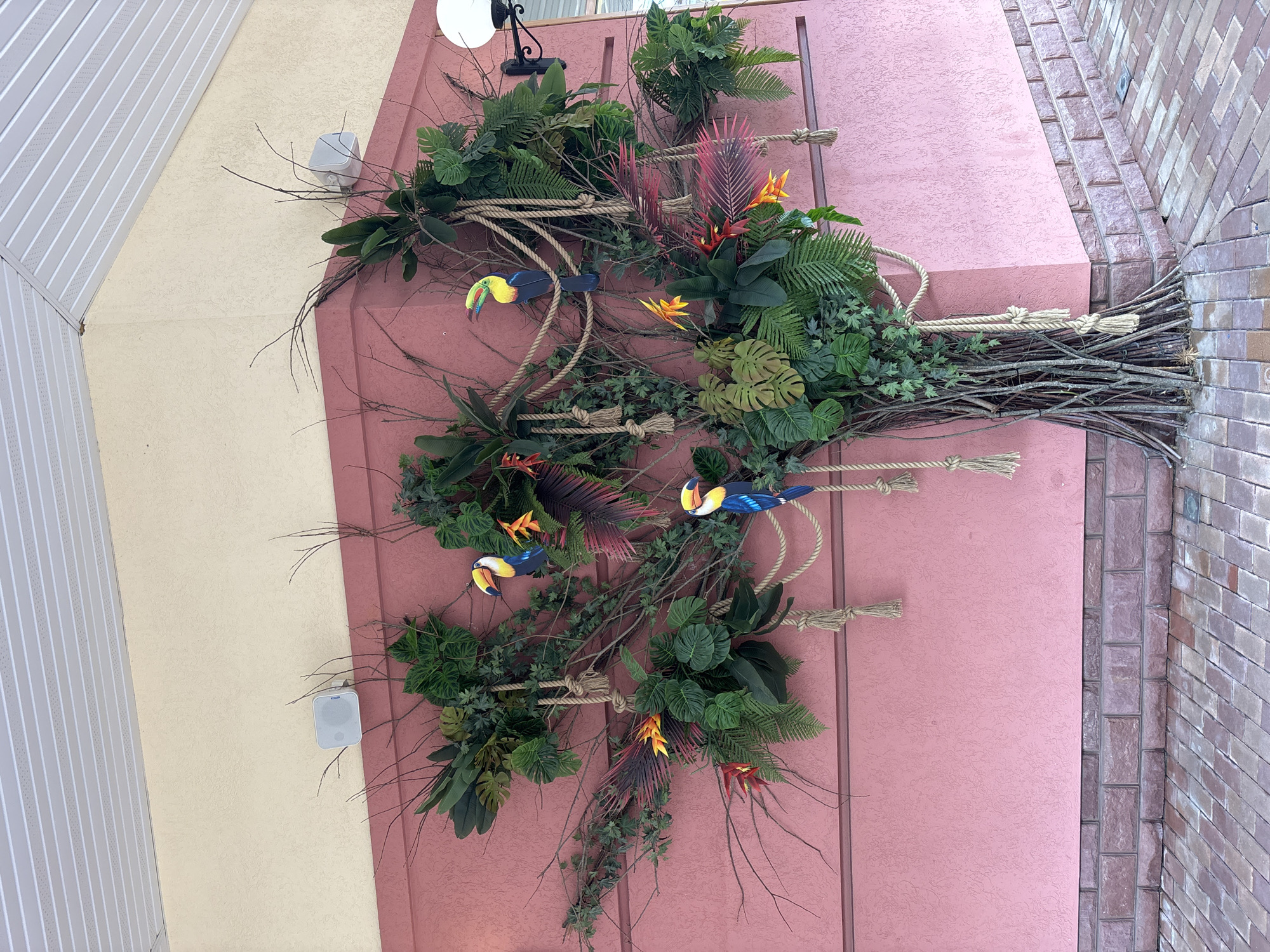 This image depicts a decorative wall installation resembling a tree, adorned with artificial tropical plants, colorful toucan figures, and thick ropes tied in knots. The installation is mounted on a textured pink wall, which is the exterior of a building. The wall is topped by a cream-colored soffit, and a white speaker is visible on the upper portion of the pink wall. To the right, a black outdoor light fixture is attached to the wall. The base of the installation rests on a brick-paved area, with the wall itself sitting atop a foundation of darker, reddish-brown bricks. The scene is likely located outdoors in an urban setting, possibly a public space or the facade of a business. The image was captured in Pokrovskoe, Russia.