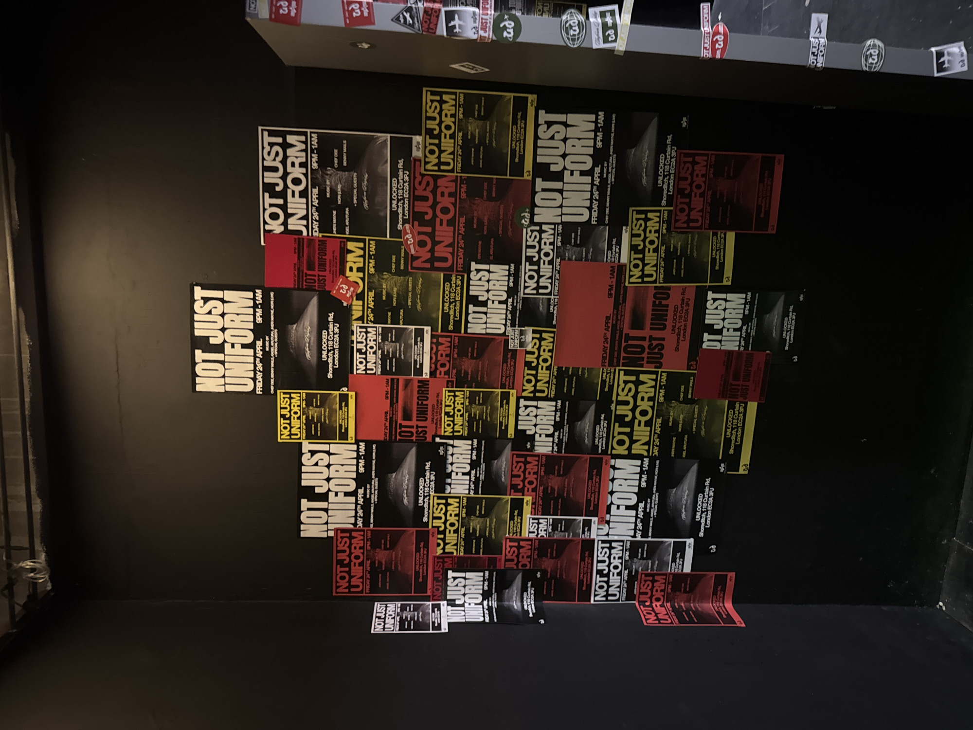 The image shows a section of a black wall covered in multiple posters for an event called "Not Just Uniform". The posters provide event details: "Unlocked Shoreditch, 118 Curtain Rd, London EC2A 3PJ", indicating the event takes place in Shoreditch, London. The posters also specify "Friday 24th April, 9PM-1AM", and list the music by Chef Dee, Benny Mails, and other artists. The posters vary in color schemes, with red, yellow, and black being prominent. Some posters are torn or peeling off the wall. A row of stickers is affixed to the top edge of the wall. The surrounding environment suggests an interior location, potentially a club or venue.