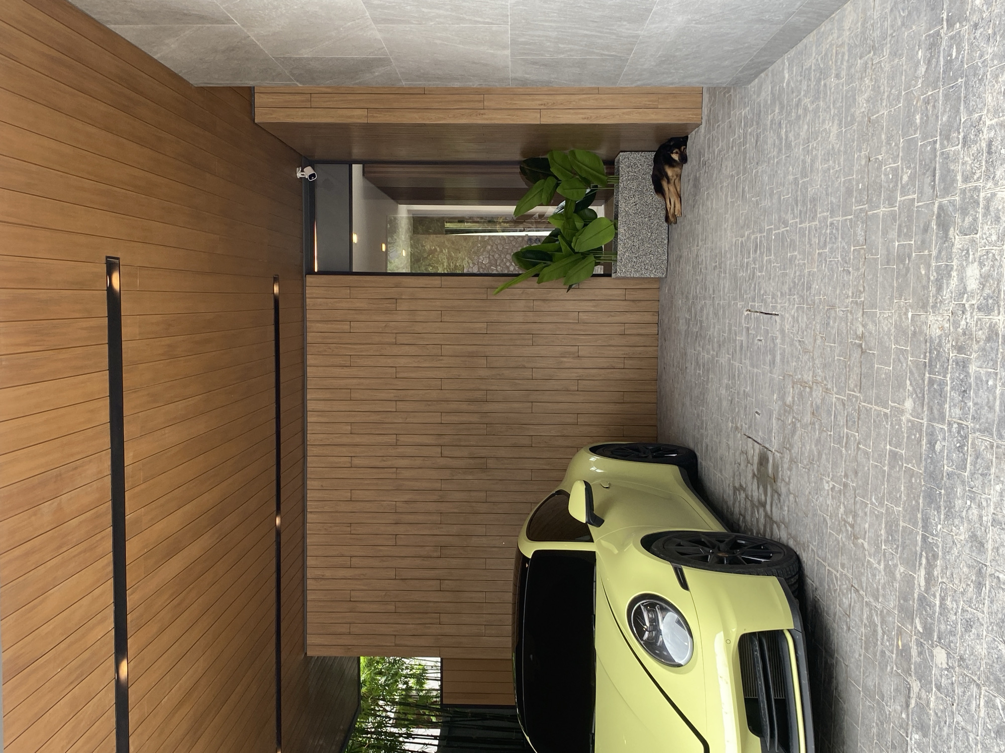 The image shows a view of a building exterior in Choeng Thale, Thailand, likely a modern residence. The architecture features a combination of materials including wooden siding, stone tile, and concrete. A pale yellow sports car is parked alongside the building on the stone tiled area. A dog is resting by a planter. Security camera are visible. A strip of greenery visible through an open air area.