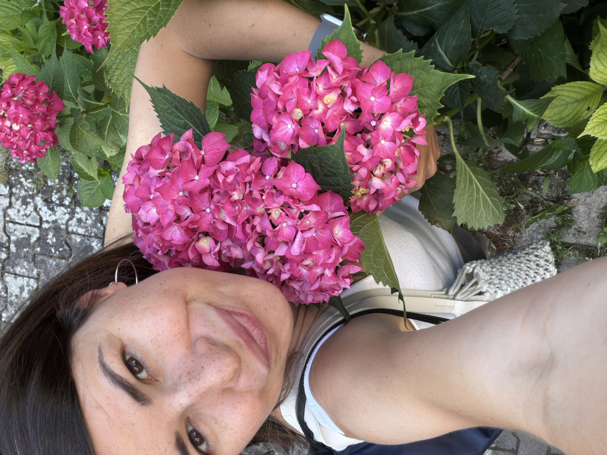 A selfie of a fair-skinned woman with brown hair and freckles, wearing a silver hoop earring, smiling upward at the camera, is the main subject. She is positioned amongst pink hydrangeas with green leaves. The background consists of stone paving, common in urban areas. The image was likely taken in Budva, Montenegro due to the prevalence of such landscaping. The subject appears to be holding the flowers. She is wearing a white top with visible straps.