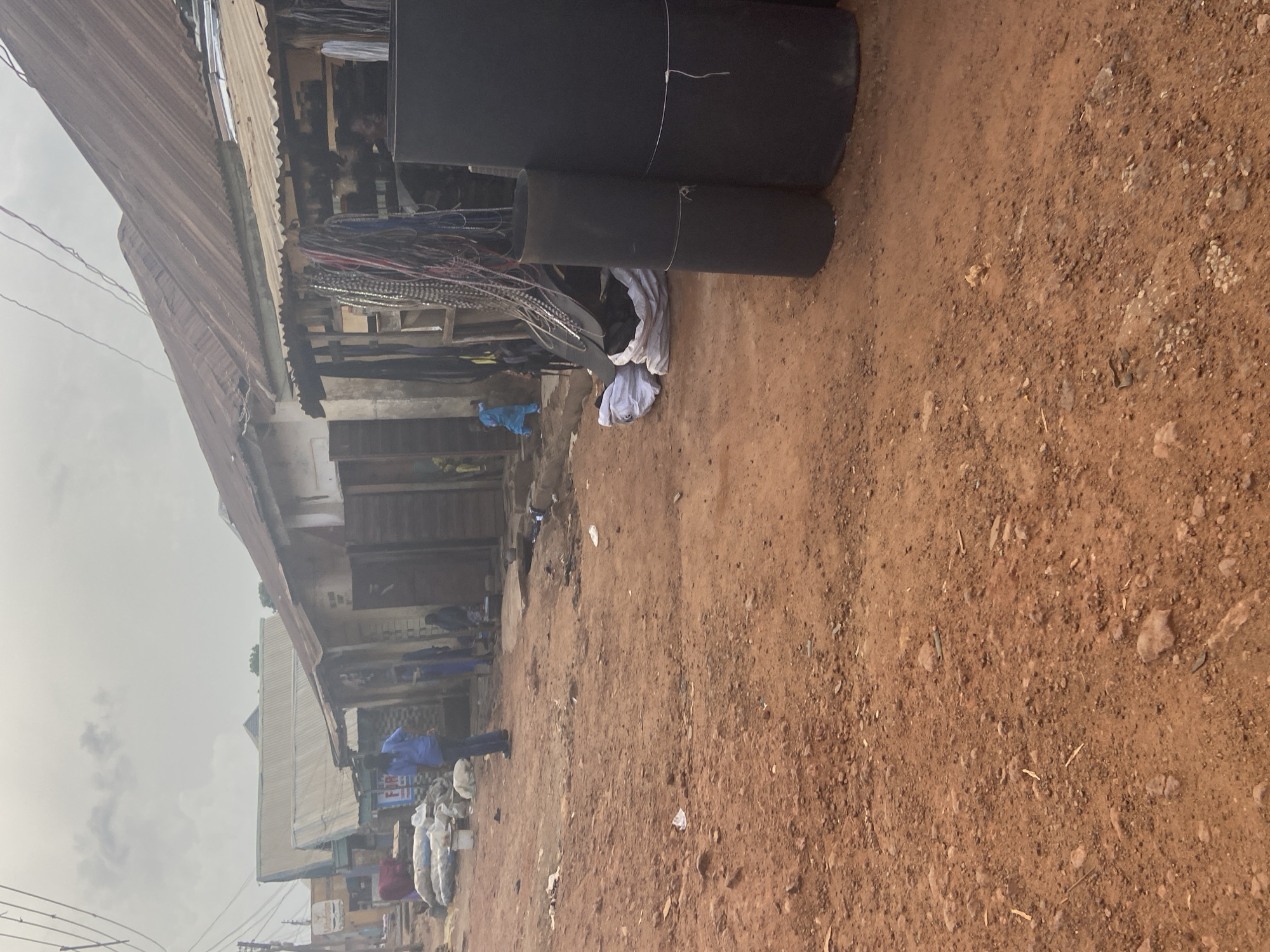 In Bukuru, Nigeria, an outdoor market scene unfolds under an overcast sky. The ground is a dusty, reddish-brown dirt, scattered with small stones and debris. Along the right side of the frame, large, black cylindrical rolls, likely material such as foam or rubber, stand upright, secured with white string. In front of them, several white bags are bundled together.
Further into the scene, a row of stalls with corrugated iron roofs line the dirt path. These stalls appear to be selling various goods, with strings of decorative items, possibly beads or chains, hanging from one stall. To the left, a man in a blue shirt and dark pants stands facing away from the camera, his back to a pile of what appear to be large white sacks.
In the background, several individuals are visible. Two people in dark clothing are near a stall on the right, one appearing to be seated. A person in a bright blue garment is walking away from the camera towards the left side of the image, in front of a building with wooden doors. The overall impression is a bustling, informal marketplace.