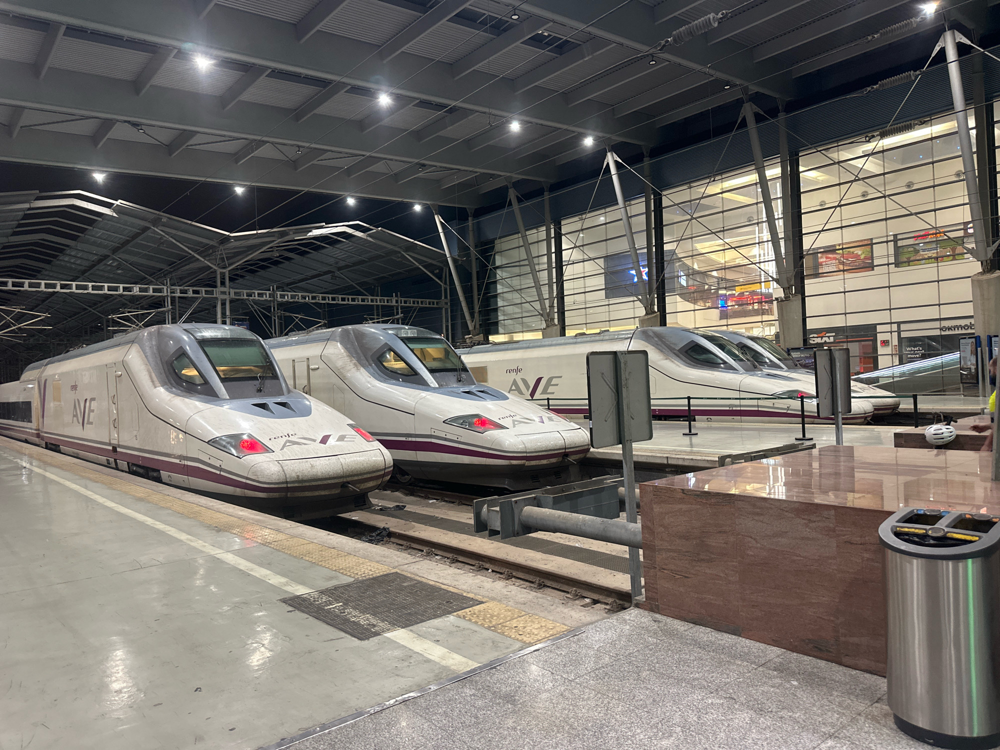 The image shows three high-speed trains, identified by the "AVE" logo and "renfe" branding, parked at a train station. The trains are white with purple accents and have a sleek, aerodynamic design. The station appears to be indoors, with a large, modern roof structure supported by metal beams and pillars. The floor is a combination of concrete and tiled sections, with train tracks running alongside the platform. In the background, there is a large glass facade that reveals the interior of what appears to be a shopping area with shops and escalators. A trash can and a marble-like structure are visible on the platform. The lighting suggests it is either evening or a dimly lit indoor space. This scene is likely at the María Zambrano train station in Málaga, Spain, which is known for its modern architecture and interconnected commercial spaces.