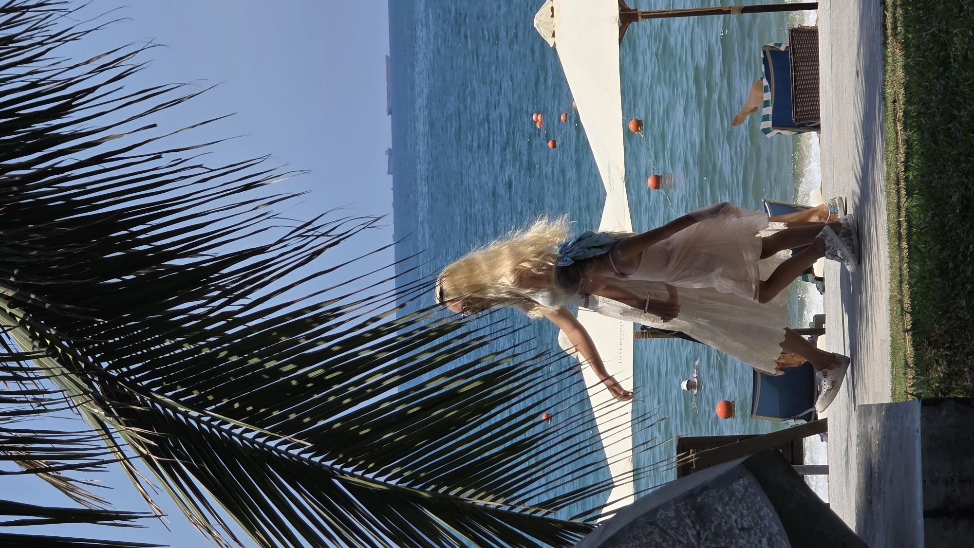 The image depicts a scene at a beachside location in Nha Trang, Vietnam. A blonde woman in a light-colored dress and sneakers stands on a paved area next to a beach. Palm fronds are visible in the foreground. The woman appears to be interacting with something near the edge of the pavement. The beach has orange buoys in the water, and beach chairs are visible in the background. The sea is calm and blue. In the distance, land or structures can be seen across the water.