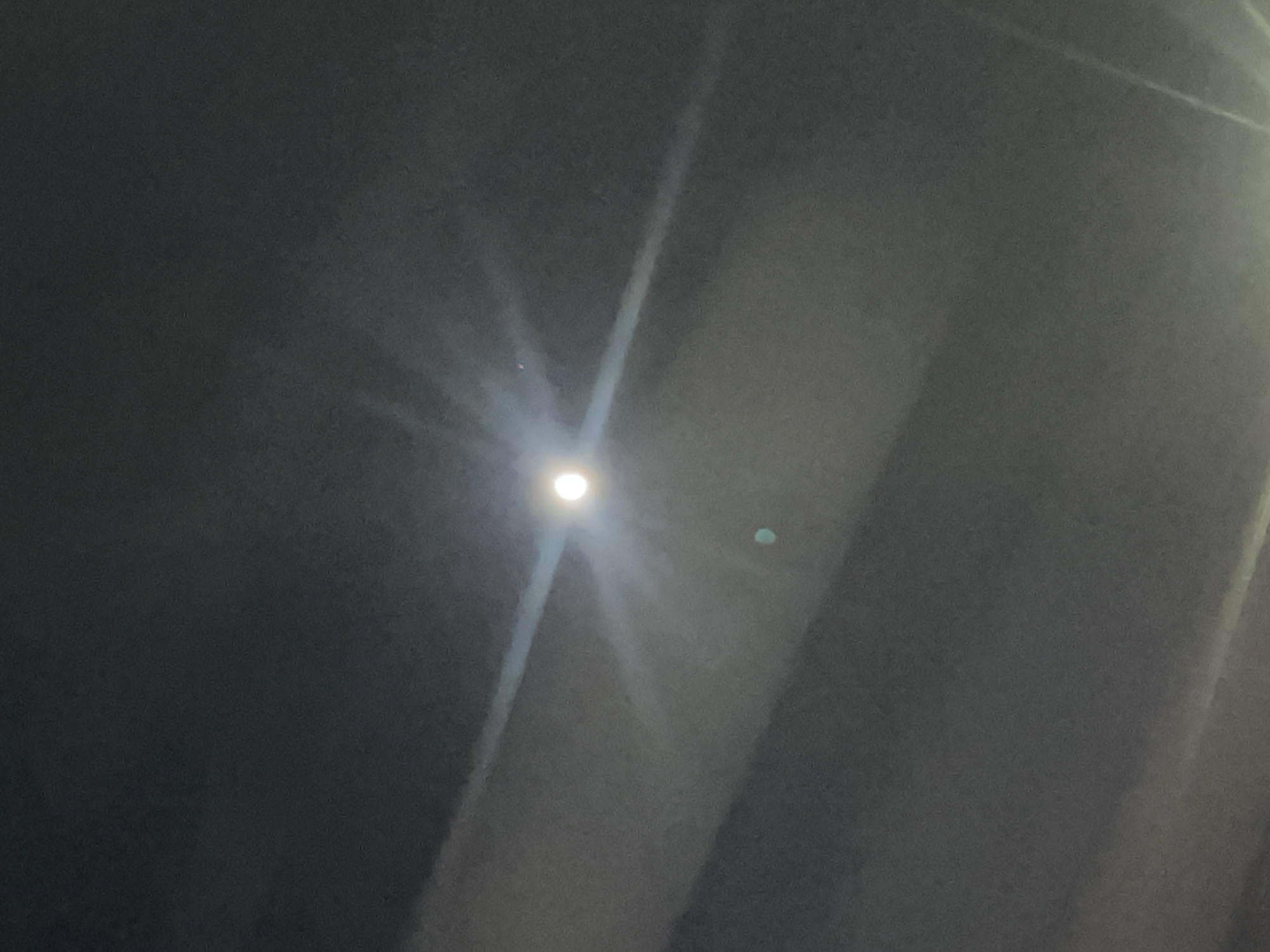 A low-angle shot of a light source, potentially the moon or a street lamp, is visible in the sky. The surrounding sky is dark and obscured by clouds or fog. A hazy beam of light extends from the light source. There are no people, subjects, objects, actions, interactions, or details of the city of Bavra, Armenia, in the image. The environment appears to be a dark, overcast night.
