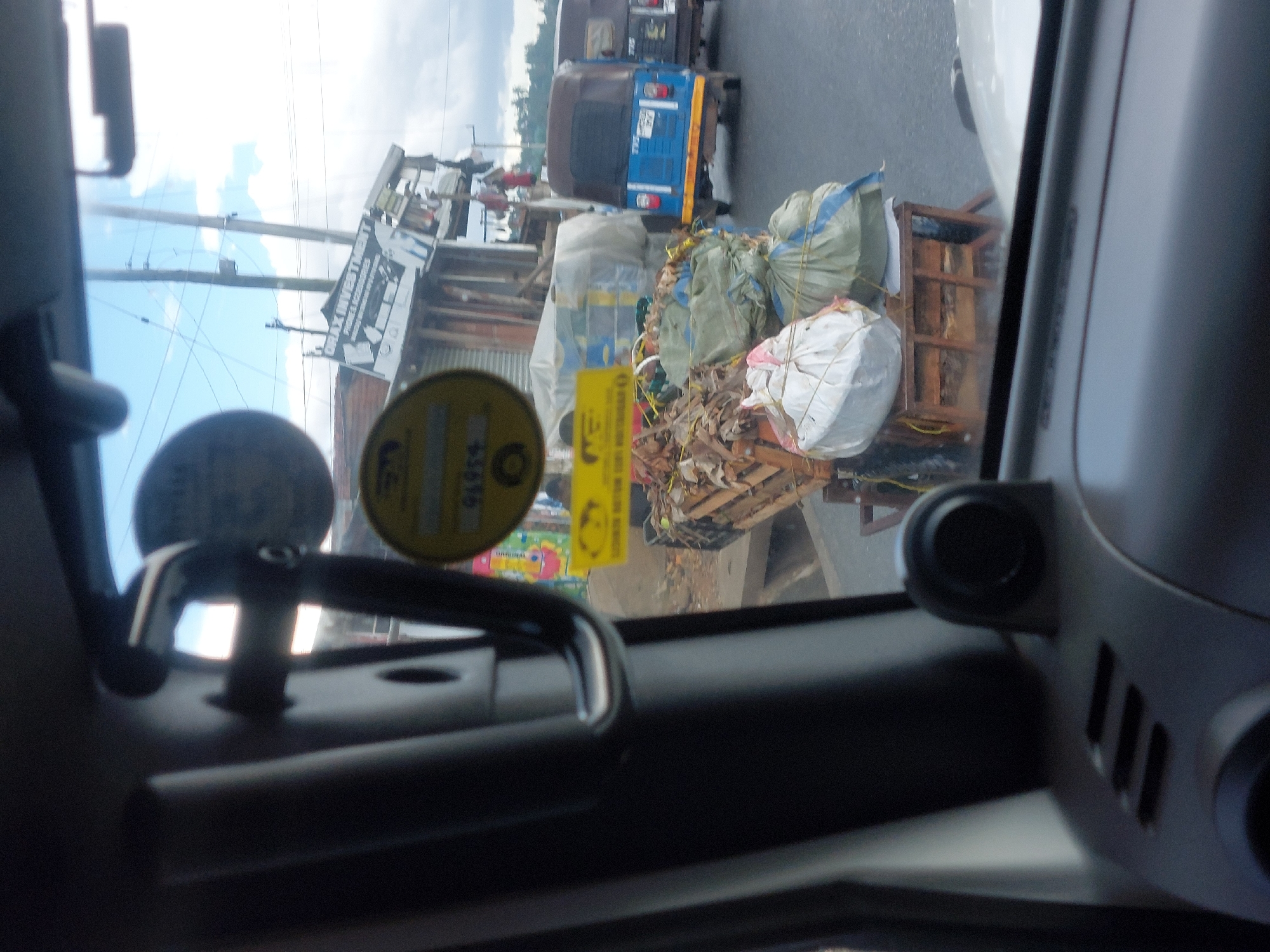 The image, taken from inside a vehicle, shows a street scene in what is likely Dar es Salaam, Tanzania. In the foreground, the interior of the vehicle is visible, including part of the dashboard and a rearview mirror. Through the windshield, the street is visible. An auto rickshaw ("Bajaji") is to the front left. A heavily overloaded cart, loaded with bags and tied-down debris and trash, is pulled by a person who is not completely visible. A building with a sign is also visible. Power lines are suspended above the street. Other vehicles are present as well. The street scene suggests an urban environment with a mix of vehicles and street-level commerce, typical of Dar es Salaam.