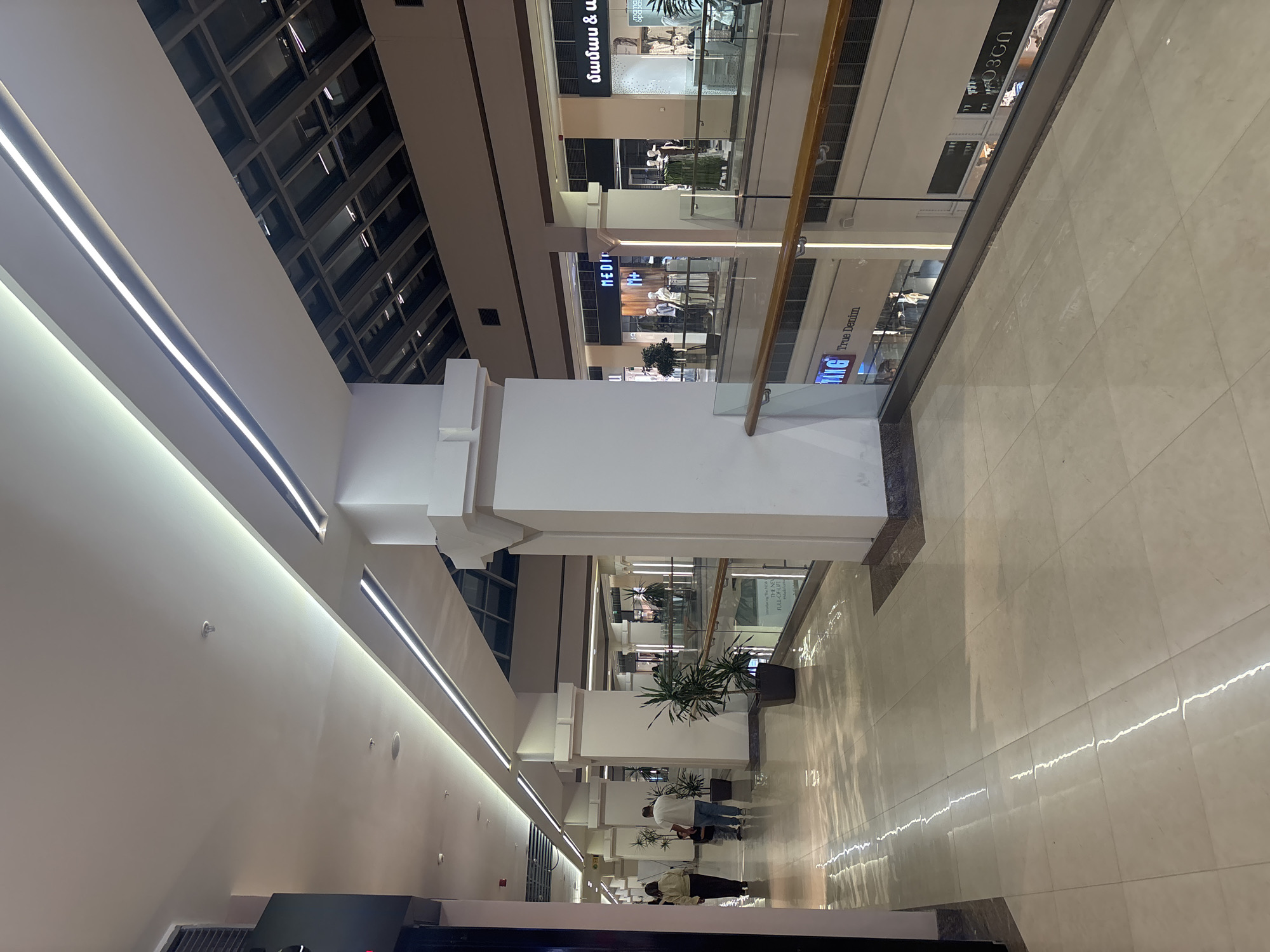 This media file captures the interior of a multi-level, modern shopping mall, likely located in Yerevan, Armenia. The perspective looks down a long corridor or atrium, featuring clean lines, a neutral color palette of white and light gray, and polished light-colored tiled floors.
The architectural design includes prominent white columns that extend upwards, supporting the upper levels. Linear LED strip lights are integrated into the ceiling and along the walls, providing bright, uniform illumination. A multi-paneled, possibly skylight or decorative ceiling structure is visible towards the top. Glass railings line the upper levels, offering views into the stores below.
Several storefronts are visible on the upper levels. One prominent sign features "MEDI" and "M+". Another display shows the text "THE NEW FULL-OF-LIFE" and a partial "MG True Denim" sign. Other text visible from the OCR includes "մամաս & և" and "O3GN", indicative of local languages. Many stores feature manikins dressed in clothing, typical of retail establishments.
A few individuals are seen walking along the ground level, suggesting general movement and activity within the public space. Potted green plants are strategically placed, adding a touch of nature to the modern environment. The polished floor reflects the bright lights, creating elongated streaks of illumination. The overall impression is one of a clean, well-lit, and contemporary commercial space. No specific time of day or weather conditions are discernible as it's an indoor setting.