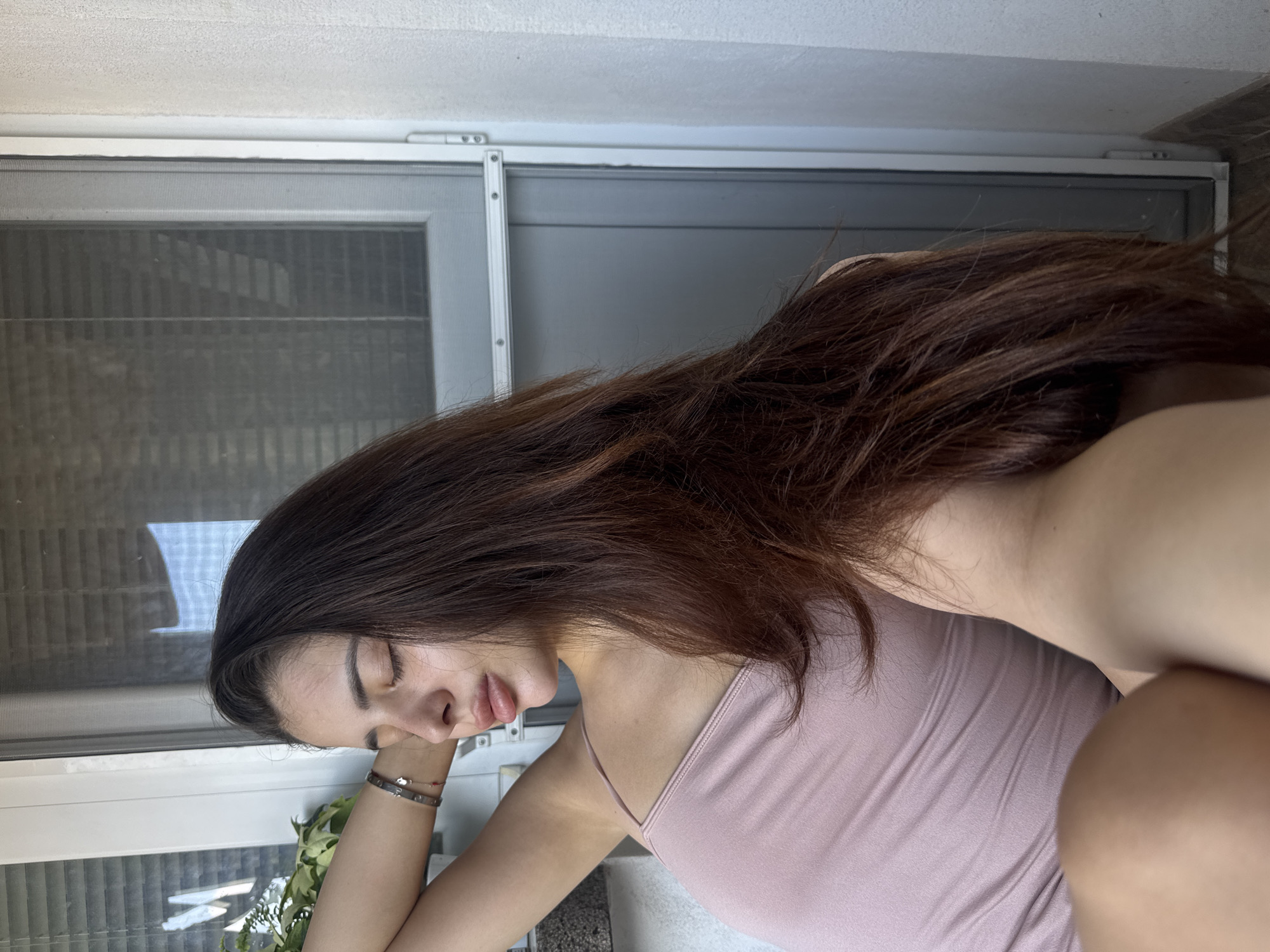 A young woman with long, dark brown hair is resting indoors, appearing calm and serene with her eyes closed, her head supported by her right hand. She is wearing a light pink or mauve spaghetti strap top and has two bracelets on her right wrist – one metallic and one a red string. The setting includes a large white-framed window with a screen, through which blurred exterior details are visible, suggesting another building or outdoor elements. A small green plant or foliage is visible near the bottom of the window. Above the window, a white, textured ceiling or wall is visible. The natural light filtering through the window indicates it is daytime. The scene is set in Plovdiv, Bulgaria. There is no visible text in the image.