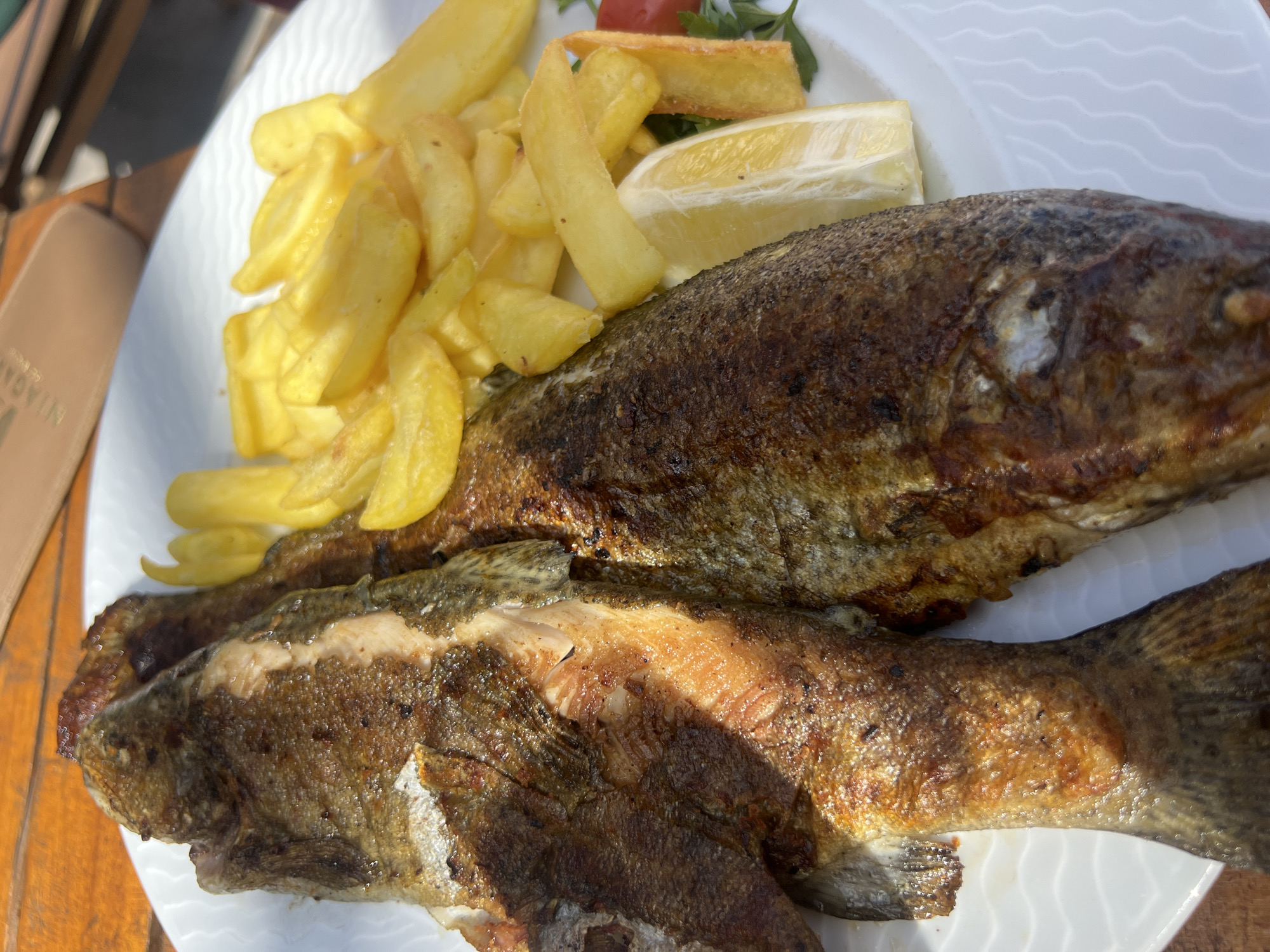 On a white plate, two whole fried fish lay next to a pile of French fries and a lemon wedge. The plate sits on a wooden table. The scene is likely in a restaurant or outdoor cafe. The location is Podgorica, Montenegro.