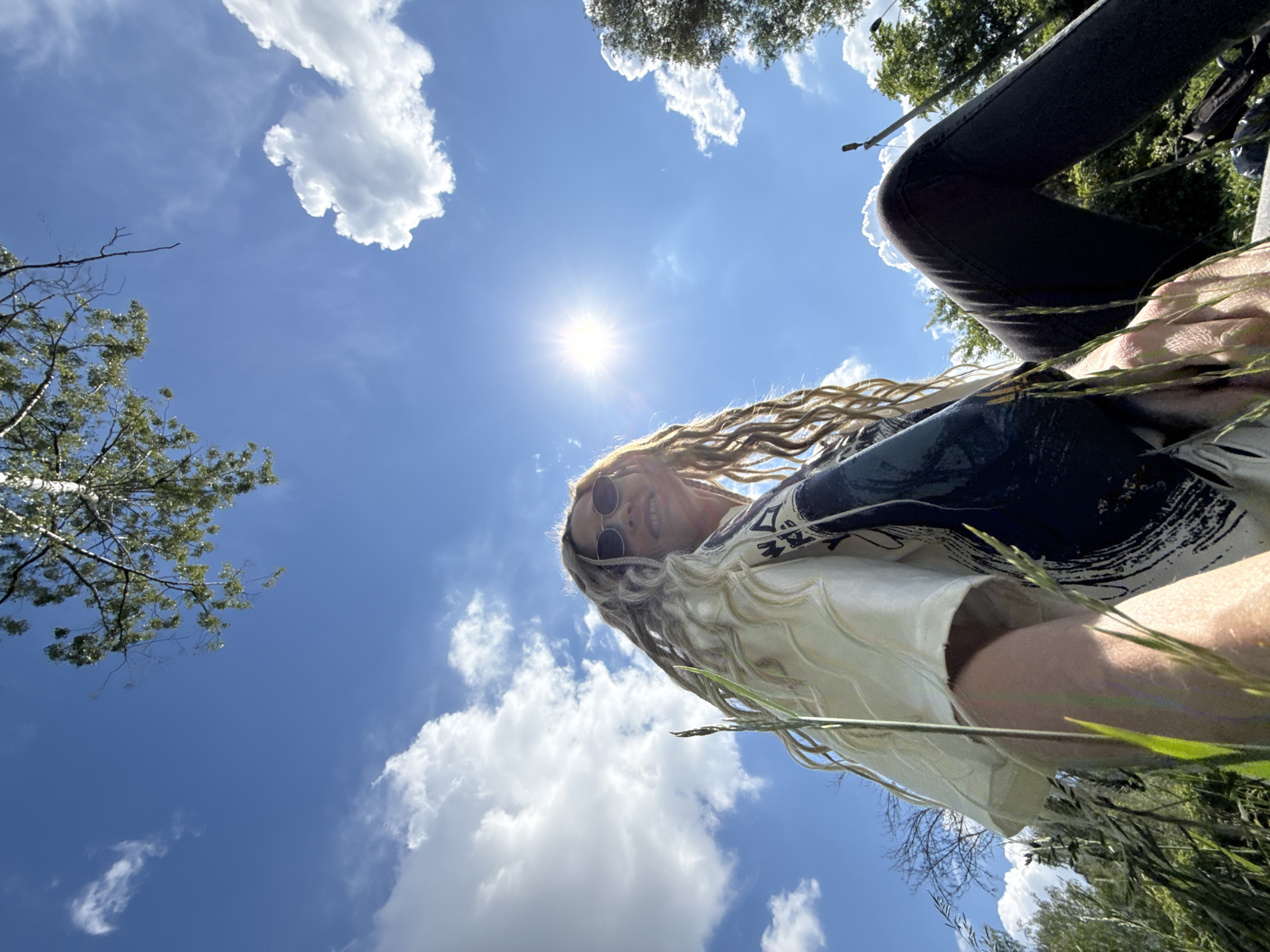A low-angle outdoor photograph captures a person from below, set against a vibrant blue sky dotted with scattered white cumulus clouds. The bright sun, visible in the upper-middle left, creates a strong starburst effect, illuminating the scene. The primary subject is a person with long, textured blonde hair (possibly dreadlocks or braids) and sunglasses, who appears to be smiling or grinning. They are wearing a light-colored top under a darker garment with abstract patterns, and dark trousers.
The image is framed by lush green grass and tall weeds in the foreground, suggesting the photo was taken from a very low perspective, possibly from the ground. Trees with green foliage are visible on the left and upper right sides, along with some bare branches, silhouetted against the bright sky. The overall impression is one of a cheerful and relaxed moment outdoors on a clear, sunny day. The setting appears natural and verdant, indicative of a park or rural landscape in Rusanovo, Russia. No visible text is present.