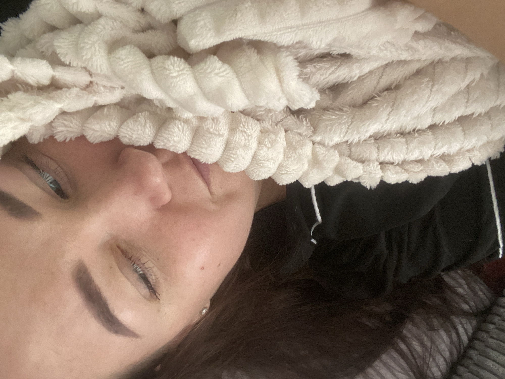 A woman with brown hair and fair skin is pictured in a close-up, partially obscured by a white, textured hair wrap. Her right eye is open, revealing a light blue iris. She is lying down, and a dark-colored hooded garment is visible behind her head. The location and city of Sochi, Russia are not identifiable in the image.