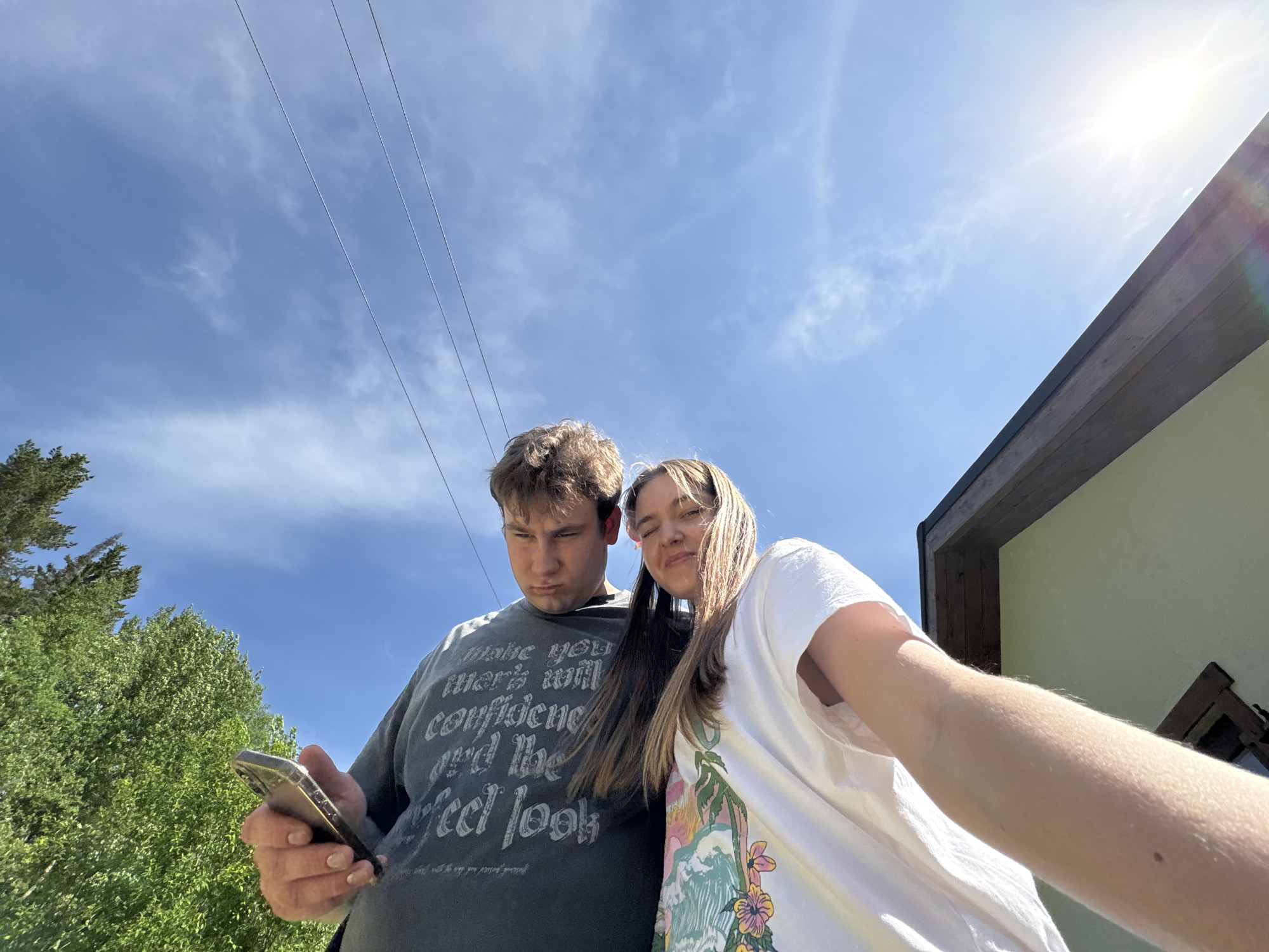 Two people, a male and a female, are in a close-up shot, likely a selfie. The male is on the left, holding a smartphone and looking down at it. He is wearing a dark gray t-shirt with printed text. The female is to his right, also looking towards the phone. She is wearing a white t-shirt with a colorful floral design. Above them is a blue sky with some clouds and power lines. Part of a green tree is visible on the left, and the corner of a building is on the right. The location is outdoors in daylight. The city or specific location within Sibir', Russia, is not discernible from this image.
