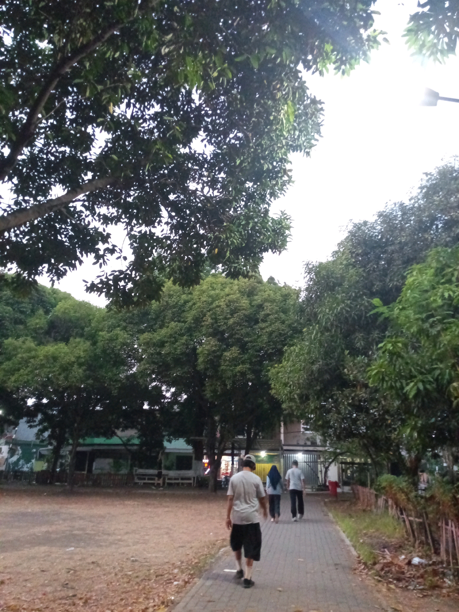 The image depicts a scene in Bekasi, Indonesia, featuring an outdoor area with trees and a paved walkway. In the foreground, a man wearing a light-colored t-shirt and dark shorts is walking away from the camera on a dirt path. Further ahead on the paved walkway, two individuals are walking in the same direction; one appears to be a woman wearing a blue headscarf and dark clothing, and the other is a person in a light-colored t-shirt and dark pants. A red object, possibly clothing, is visible on the ground near the walkway. The background shows lush green trees, some buildings with visible awnings, and a street lamp. The sky is bright, suggesting it is daytime, possibly late afternoon or early morning due to the diffused light. The overall environment appears to be a park or a public recreational space within the city.