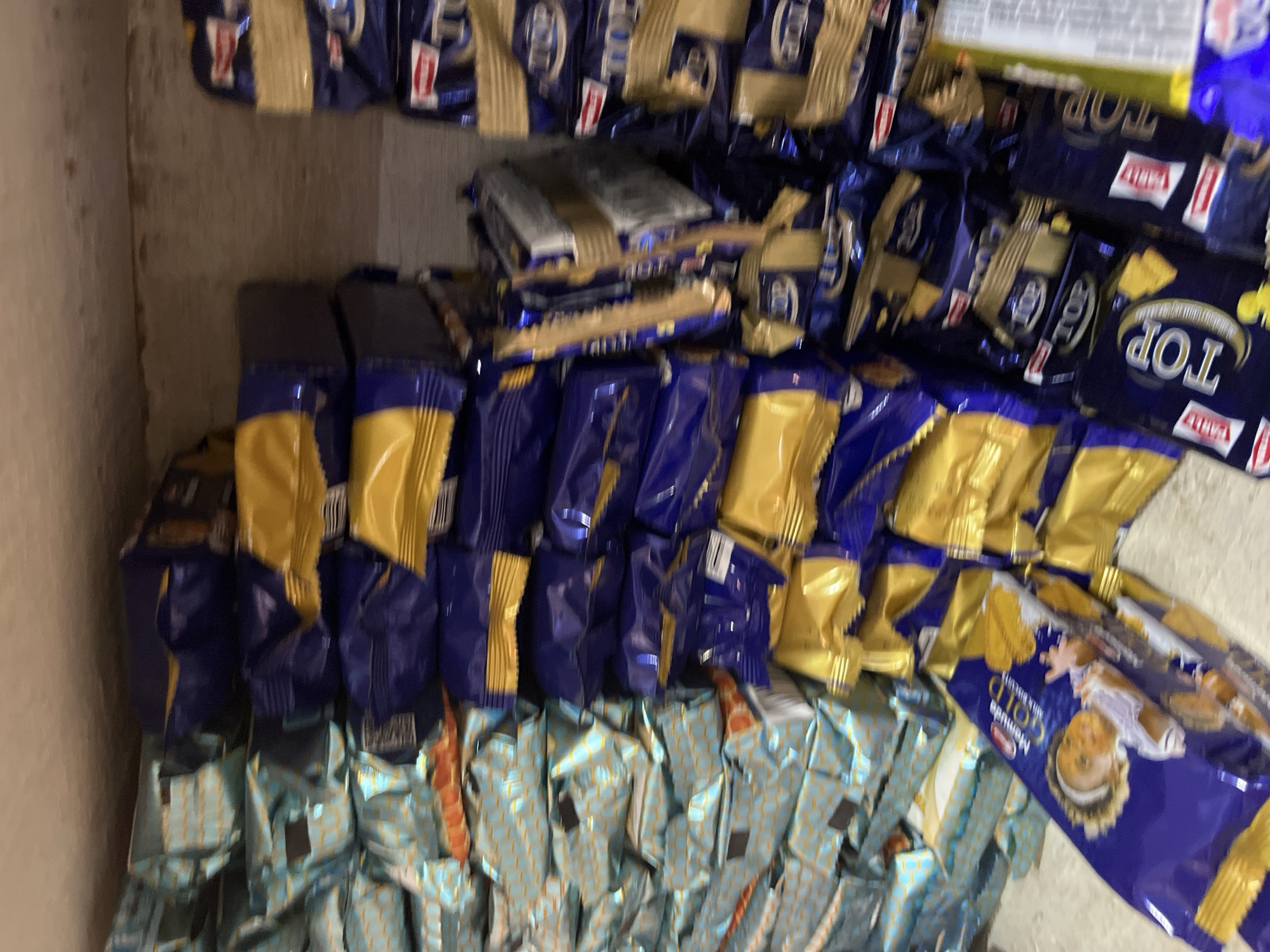 The image shows a large stack of packaged biscuits. The packaging is predominantly blue and gold, with some packages featuring a light blue and gold pattern. Several packages have the word "TOP" printed on them, along with an image of a crescent moon and stars. One package, in the foreground and angled to the viewer's left, clearly displays "McVities GOLD MILK BISCUIT" with a picture of a baby. The biscuits are packed tightly together, suggesting a storage area or a retail environment. The overall impression is one of abundance and bulk storage of confectionery products. The image does not provide any information or context that would allow for the determination of its location in the city of Bukuru, Nigeria.