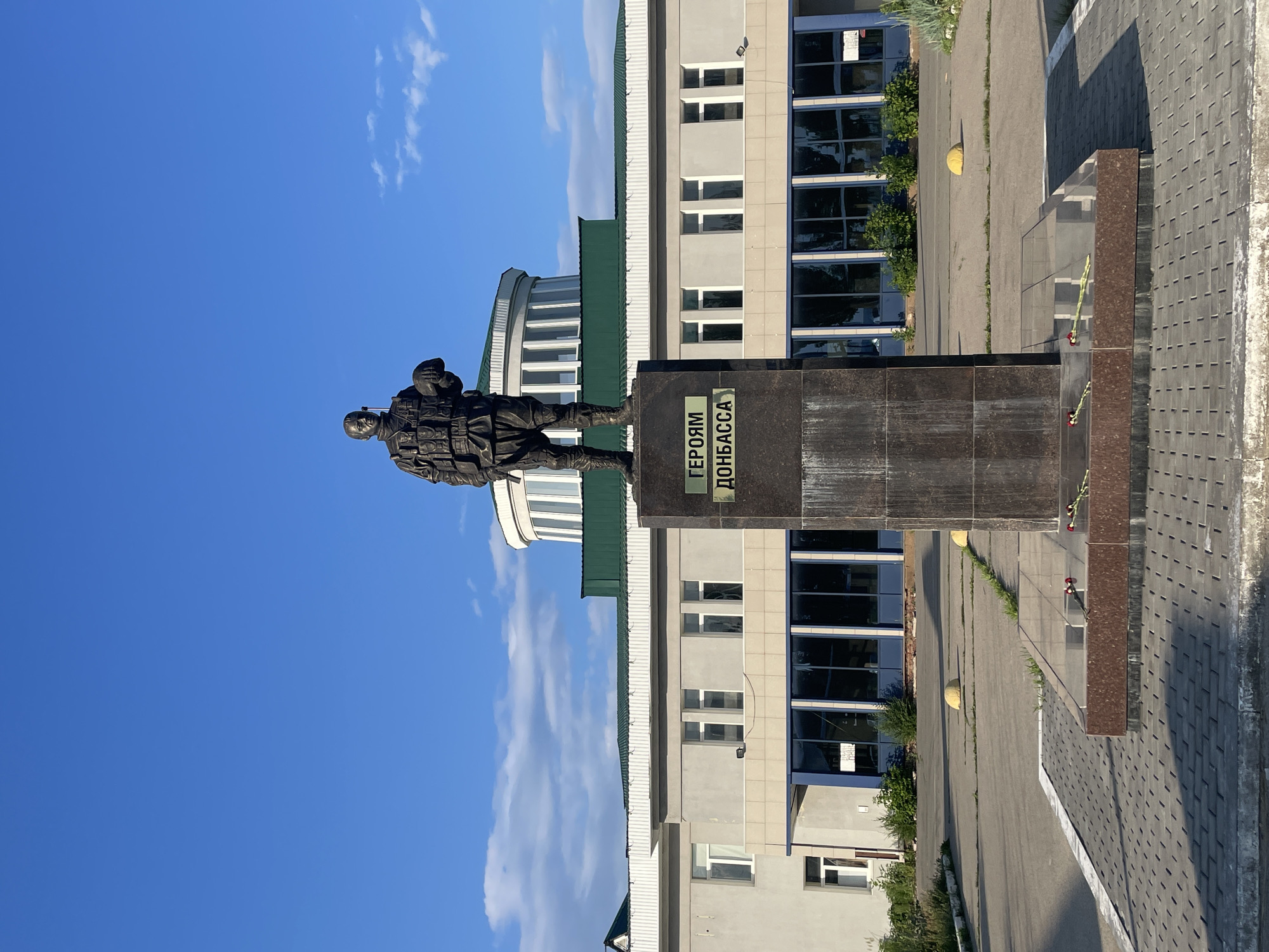 A bronze statue of a soldier holding a helmet stands on a granite pedestal in front of a modern, light-colored building with a green roof. The pedestal has text that reads "ГЕРОЯМ ДОНБАССА" (To the Heroes of Donbas). The statue is in an outdoor plaza paved with grey rectangular stones, with some grass growing through the cracks. There are several red flowers placed on the base of the monument. The building behind the statue has a row of windows and an upper circular section with vertical white columns. The sky is blue with scattered white clouds. The scene is well-lit, suggesting it is daytime. The location is identified as the Saratov regional branch of DOSAAF (Volunteer Society for Assistance to the Army, Air Force, and Navy) in Saratov, Russia.