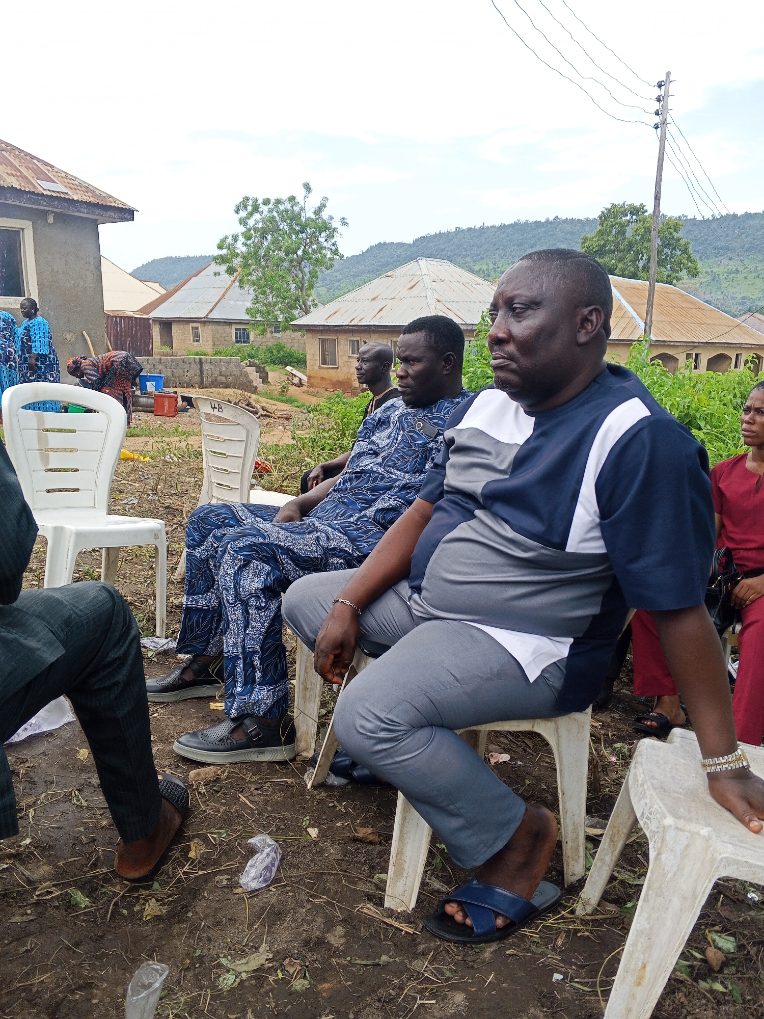 In Lokoja, Nigeria, several individuals are seated outdoors on plastic chairs, suggesting a community gathering or event. The ground is uneven and appears to be dirt, with scattered dry leaves and some plastic debris. In the background, traditional Nigerian houses with corrugated metal roofs are visible, nestled among vegetation and a backdrop of rolling hills. A power pole with visible wires stands to the right, indicating the presence of electricity infrastructure. The people are dressed in a mix of traditional and contemporary clothing, with some wearing vibrant patterned fabrics and others in more subdued colors. The overall atmosphere seems relaxed, with some individuals looking towards the camera and others engaged in conversation or observation. The lighting suggests it is daytime, possibly overcast.