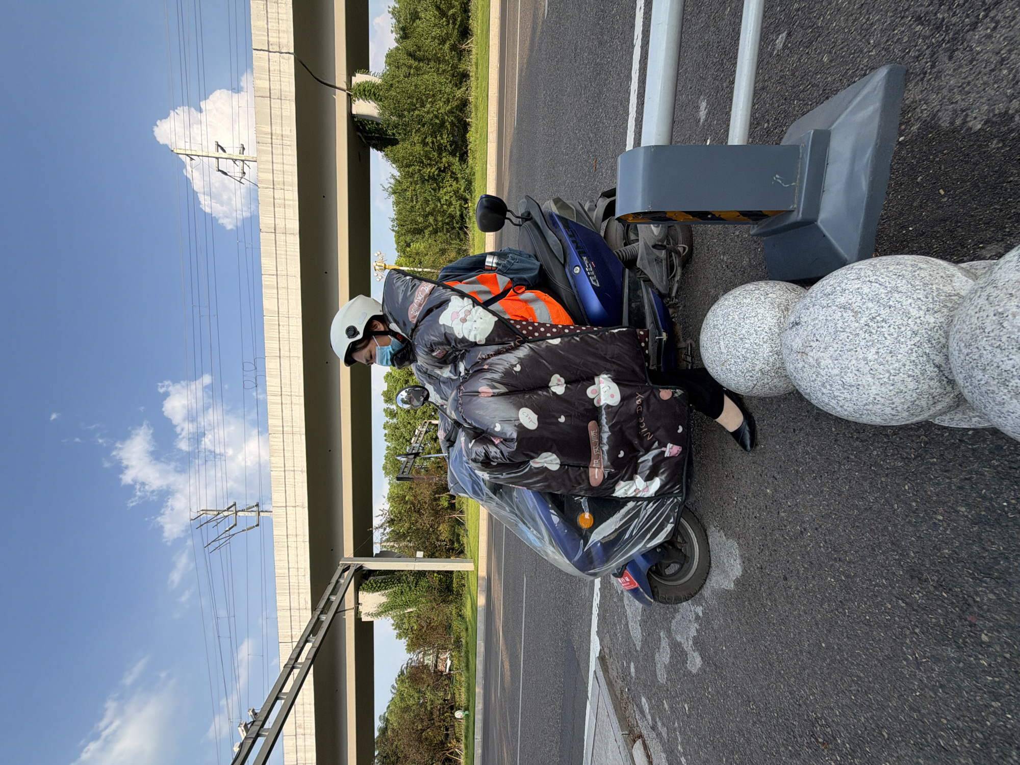 An adult, wearing a white helmet and a blue medical mask, is seated on a dark blue electric scooter. The person is wrapped in a large, black puffer coat with a pattern of white cartoon animals and the words "Nice Day." The scooter is covered with a clear plastic sheet. The scooter is parked on the side of an asphalt road, next to a grey concrete barrier with horizontal white and yellow stripes. In the foreground, three large, spherical granite bollards are partially visible. In the background, a multi-lane road is visible, separated by a white line. Overhead, an elevated concrete structure, likely part of a highway or railway, with overhead power lines, spans across the scene. Lush green trees and vegetation line the right side of the road. The sky is blue with scattered white clouds, suggesting daytime. The scene appears to be on a road in or near the city of Jiangjin, China.