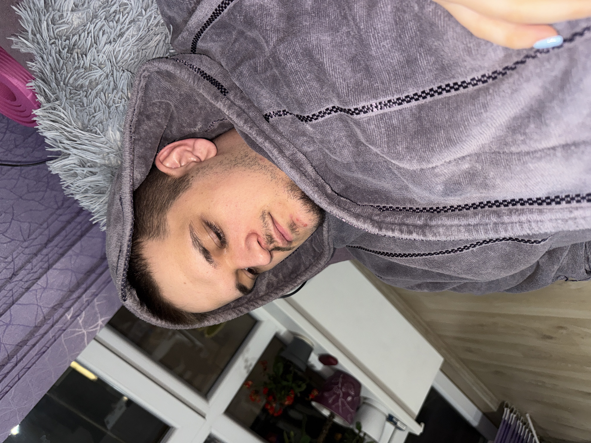 A person is lying on a bed covered with a grey furry blanket and a lavender patterned sheet, wearing a grey hoodie. The person's head is partially covered by the hoodie. A pink foam roller is visible near the blanket. A window is visible in the background, revealing a potted plant with red flowers and a white surface. The exact location in Plovdiv, Bulgaria is not discernible from the image.