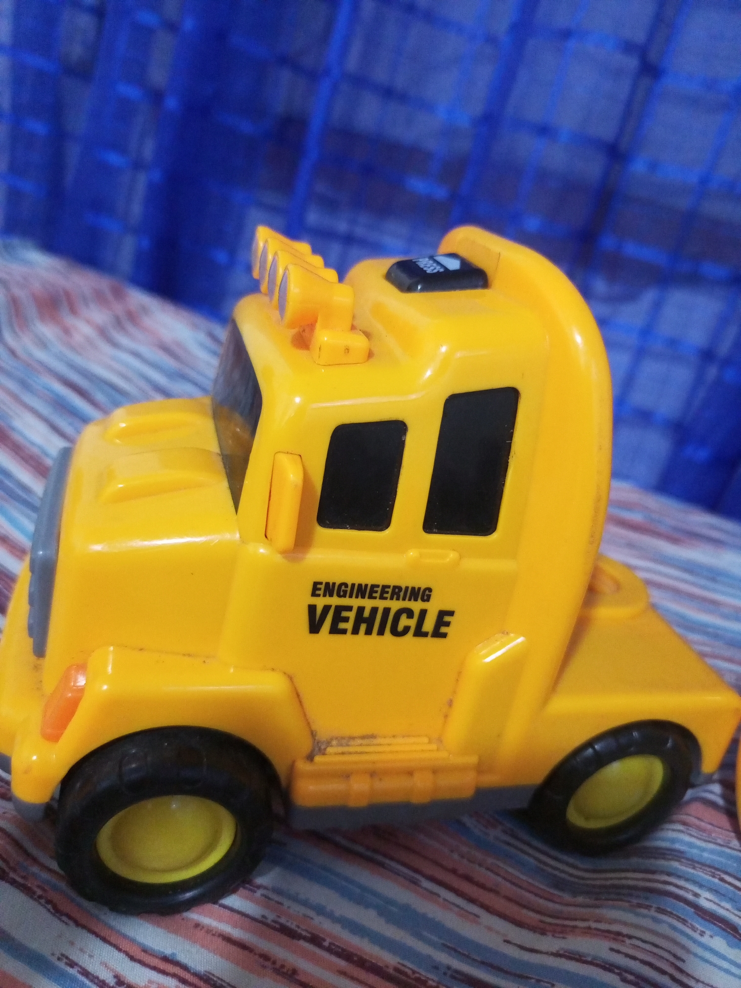 A yellow toy truck, labeled "ENGINEERING VEHICLE," sits on a surface with striped fabric in the foreground and a blue, gridded background. The truck has black wheels with yellow rims and orange indicator lights on the front. It also features a row of yellow lights on the roof and a black button on top. The scene appears to be indoors, with the fabric and blue background suggesting a domestic setting. There is no indication of the location being La Tagua, Venezuela.