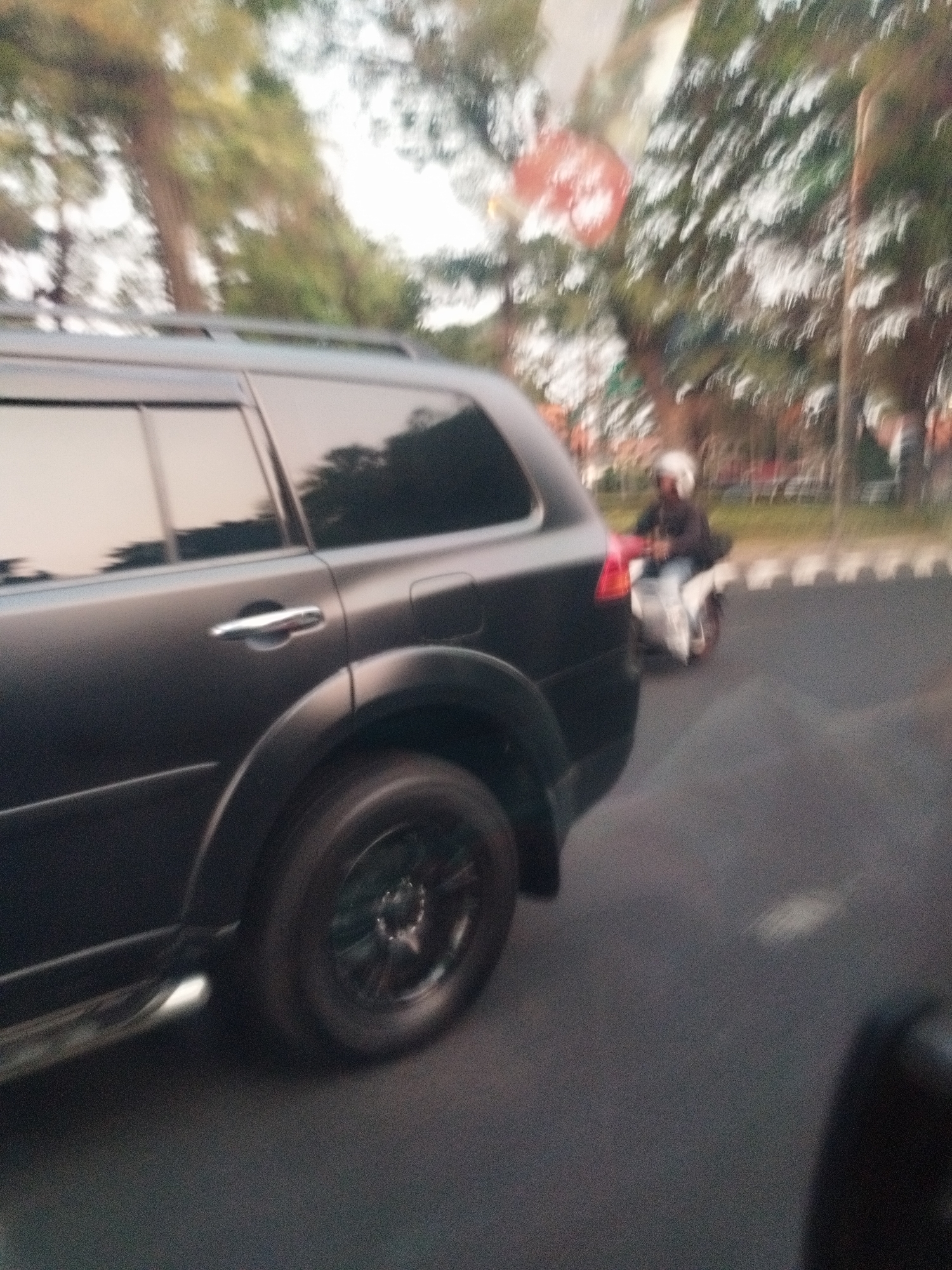 The image shows a side view of a dark-colored SUV parked on the side of a road. The SUV has tinted windows and a roof rack. A person on a white scooter is visible in the background, moving in the same direction as the SUV. The scooter is white with red accents and the rider is wearing a white helmet and dark clothing. The background shows trees, a sidewalk, and a street with road markings. The overall scene appears to be an outdoor environment, possibly a park or a residential area. The location is identified as Surabaya, Indonesia.