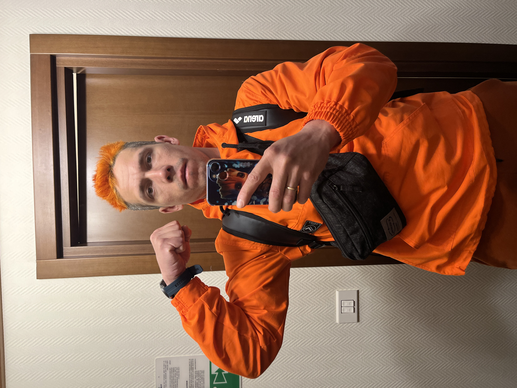 A man in an orange jacket and orange hair is taking a selfie in a room. He is wearing a black bag across his chest. The room has a wooden door, a light switch on the wall, and a textured wallpaper. The man is holding up his left fist and looking at the phone. The phone case appears to have an image of Groot from Guardians of the Galaxy. It is impossible to confirm the location is Moscow, Russia, without additional information.