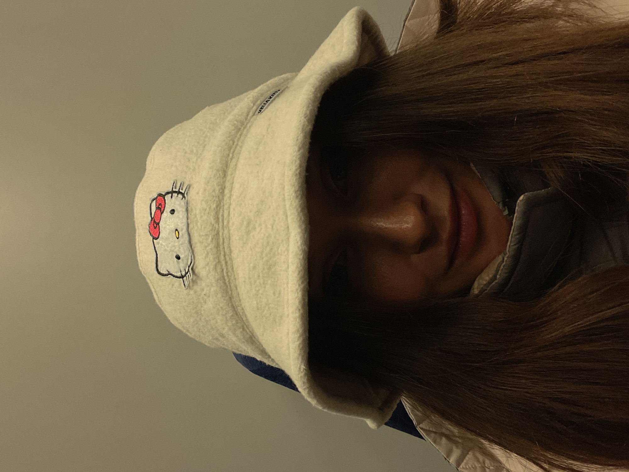 A person with long brown hair is wearing a white bucket hat with a Hello Kitty patch. The person is smiling slightly. The background is a light gray wall. There is no indication of Sochi, Russia in the image.