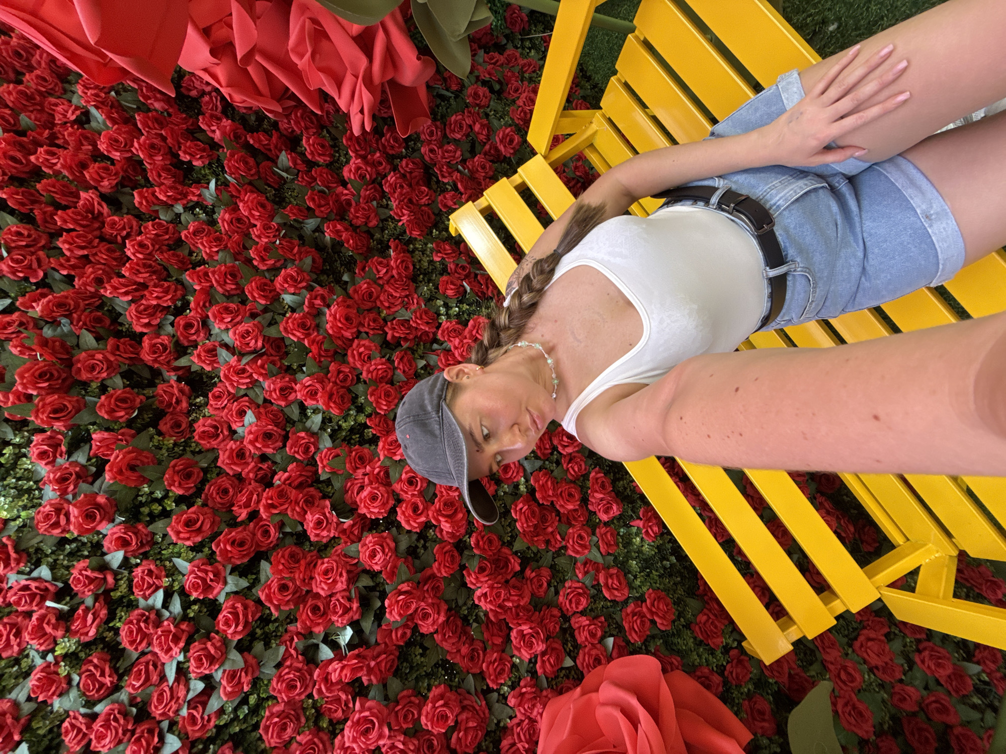 A young woman is captured in a selfie taken in front of a wall of red roses. She is sitting on a yellow bench and wearing a white tank top, denim shorts, and a baseball cap. The backdrop is a dense arrangement of artificial red roses with green foliage. To the left of the woman, a large, oversized red rose decoration is visible. The scene appears to be an outdoor setting, possibly a public space or event area in Yiwu, China.