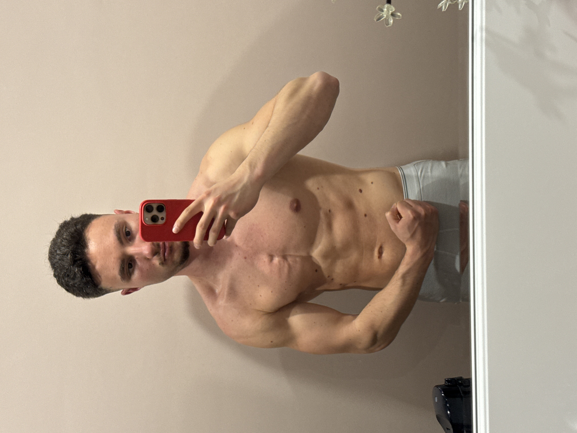A young adult male is captured taking a mirror selfie indoors, posing to display his physique. He is shirtless, wearing gray boxer briefs, and holding a red smartphone with his right hand, which partially obscures the lower part of his face. His left arm is flexed, showcasing his bicep, and his core muscles are engaged, revealing definition in his abs and chest. He has short, dark hair and stubble, and his expression is neutral.
The background reflected in the mirror is a plain, light beige wall, suggesting a private indoor setting such as a bedroom or bathroom. A vertical white frame or door jamb is visible on the right side of the mirror's reflection. Several small moles are scattered across the man's torso and arms. In the upper right corner of the mirror, a reflection of a translucent decorative object, possibly a light fixture or wall art, can be seen. A dark object, appearing to be an electronic device, is visible at the bottom right. The image is presented rotated. While not visually discernible from the image itself, the location is specified as Sofia, Bulgaria.