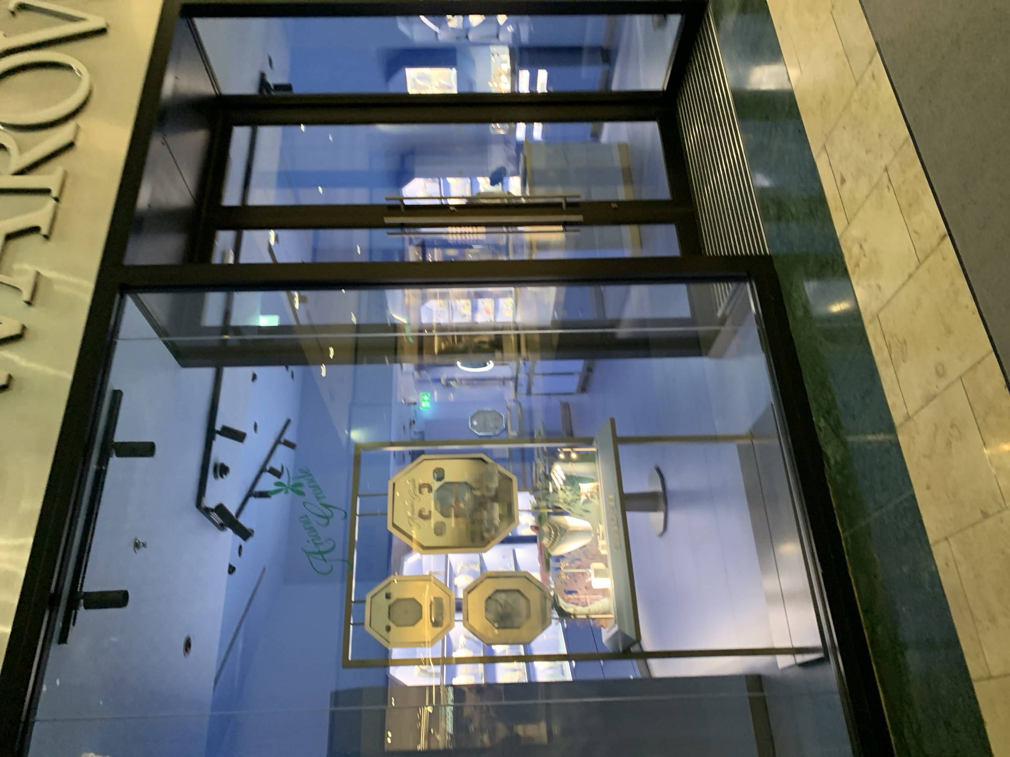The image shows the facade of a retail store, likely a jewelry or gift shop, as seen through its glass storefront. A sign on the left reads, in part, "Aroma Grune," possibly indicating the store's name. Inside the store, displays of small, gold-colored objects are visible. The store is located in Dublin 16, Ireland. The architectural details suggest a modern shopping center or mall setting. There are no people visible in the image.