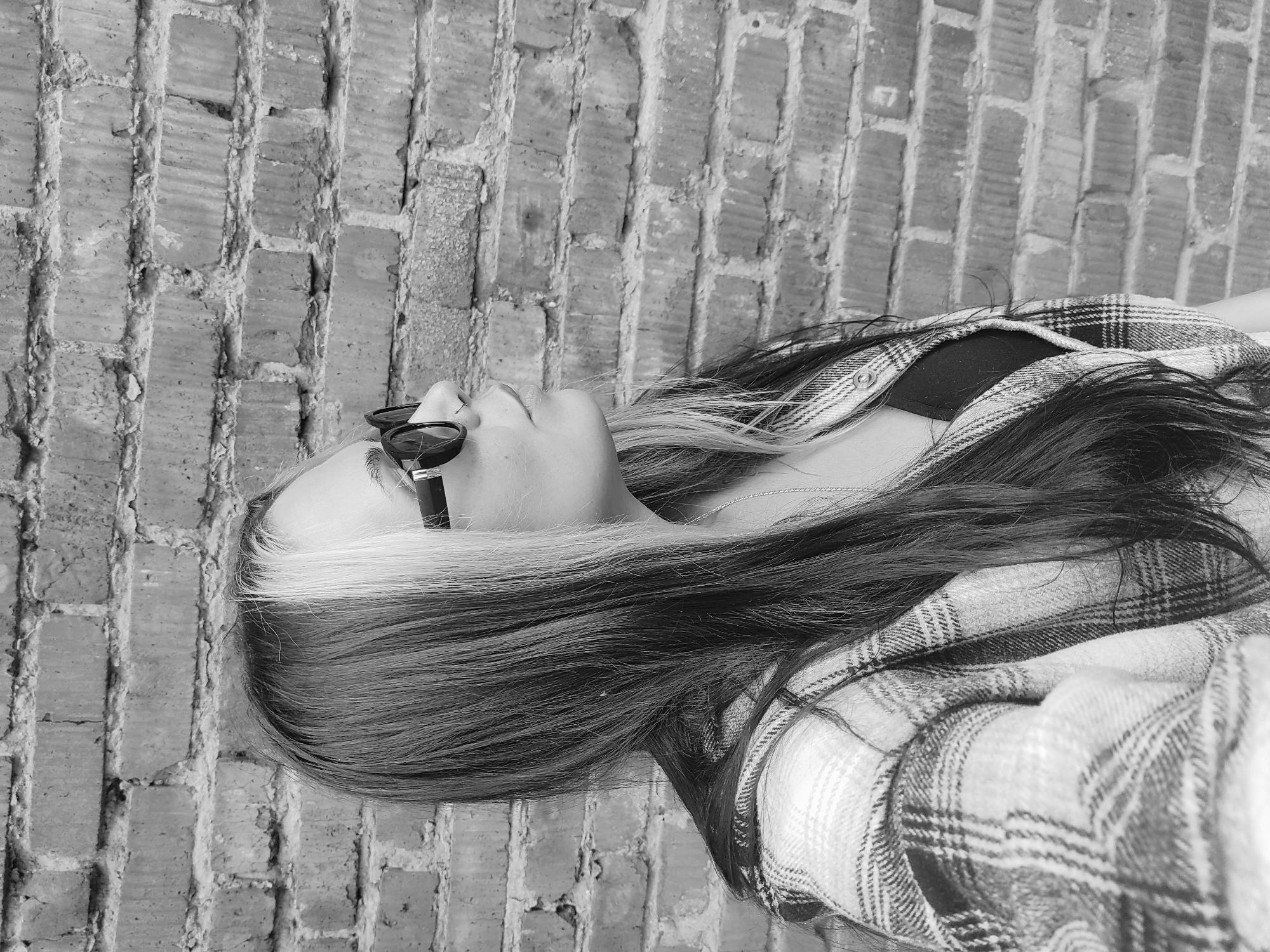 A monochromatic, high-angle shot depicts a young person, likely female, lying horizontally with their head tilted upwards against a brick wall in Chaikovsky, Russia. The individual is positioned so their head and upper torso are visible, with their long hair spread out on the brick surface.
The subject wears dark sunglasses, obscuring their eyes. A dark top, possibly a bralette or tank top, is visible beneath an open, long-sleeved plaid shirt or jacket with a distinctive pattern of light and dark squares. A thin, silver-colored necklace rests around their neck. Their hair is a prominent feature, primarily dark, with a striking, wide streak of light-colored or blonde hair visible near the front of their head and along their face.
The background consists entirely of an old, weathered brick wall, characterized by irregular brick patterns and rough mortar lines, suggesting an aged structure. The lighting, rendered in black and white, appears even, indicating daylight, possibly under an overcast sky or indirect light, as there are no harsh shadows. The person's relaxed pose, looking upwards, conveys a sense of contemplation or calm. No text is visible in the image.