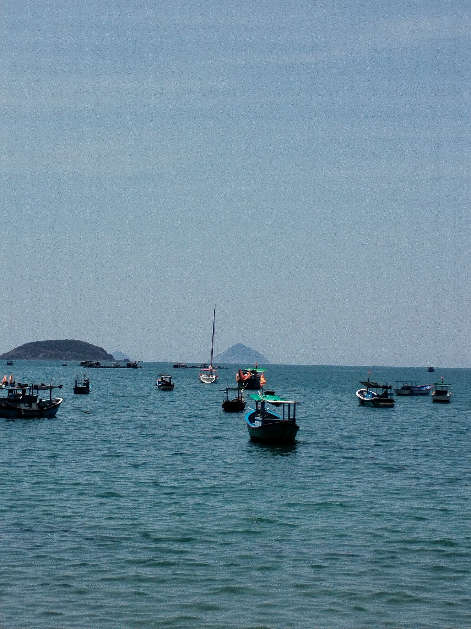 The image depicts a serene seascape in Bac Nha Trang Ward, Vietnam. The tranquil, deep-blue sea is populated with various boats, including fishing vessels and a sailboat, scattered across the water's surface. In the background, a series of islands or land formations create a layered horizon against the clear sky. The scene is quiet and peaceful, indicative of a typical maritime setting in this coastal region.