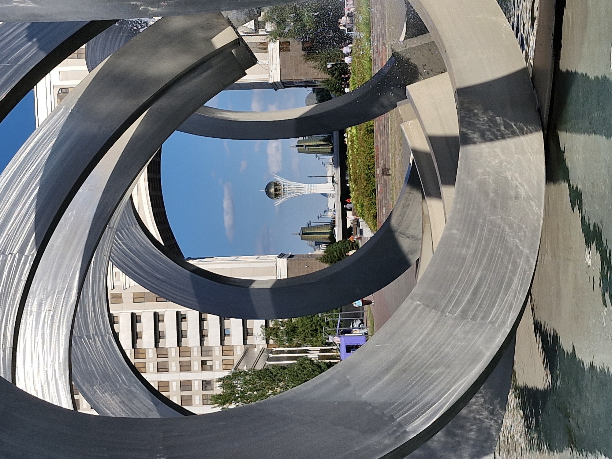 This image captures a view through a large, abstract metal sculpture, likely located in Astana, Kazakhstan. The sculpture, composed of swirling, curved metallic elements, dominates the foreground and reflects in the shallow water below.
Through the openings of the sculpture, the cityscape of Astana is visible. A prominent landmark, the Bayterek Tower with its golden sphere at the top, stands in the background, a distinctive symbol of the city. Modern administrative buildings with multiple windows and light-colored facades are also present.
In the middle ground, there's a well-maintained green area with hedges and trees. Several people are scattered throughout the scene: a few individuals walk along a paved path, while a group is seated on benches in the distance, possibly enjoying the urban park. One person is seen near a small purple structure. The sky is a clear blue with a few scattered clouds, indicating fair weather. The overall scene suggests a public space in Astana, designed to be both an artistic showcase and a recreational area.