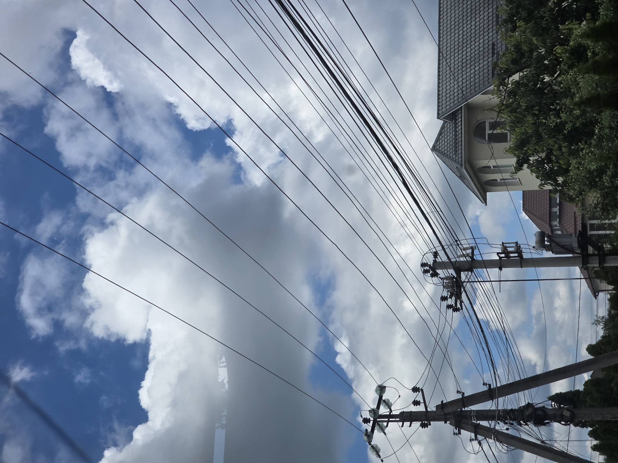 The image depicts a street scene in Petropavlivska Borshchahivka, Ukraine, characterized by a cloudy sky and overhead power lines. In the foreground and midground, two concrete utility poles support a complex network of electrical wires. The pole on the left is a triangular structure, while the one on the right is a single, taller pole. Several houses are visible in the background, with tiled roofs and architectural details that suggest residential buildings. Lush green trees form a dense canopy in front of the houses, partially obscuring them. The sky is a mix of blue patches and large, fluffy white clouds, indicating a dynamic weather pattern. There are no people or vehicles present in the frame. The overall impression is of a typical suburban or semi-urban environment under a partly cloudy sky.