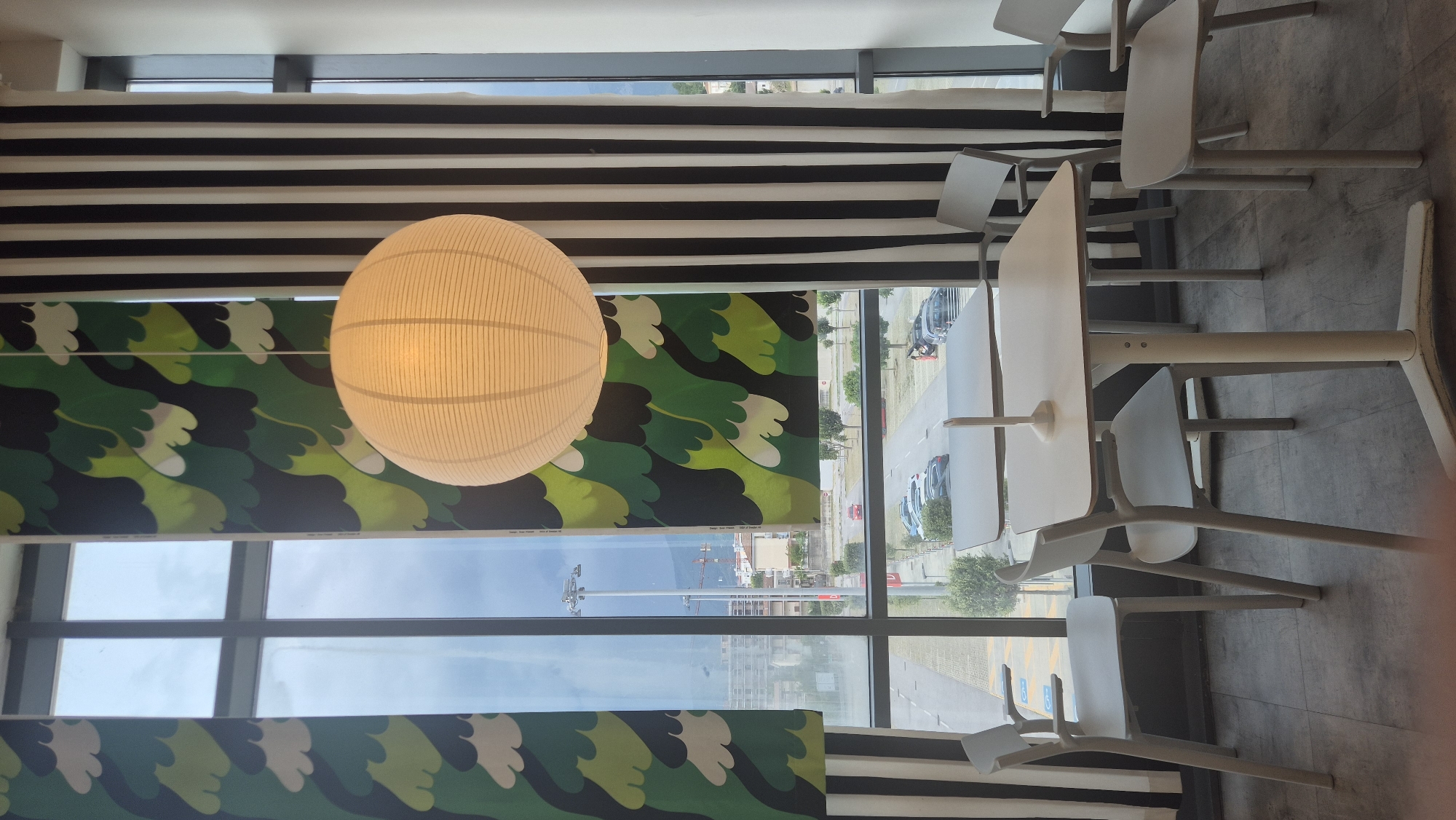 The image shows a seating area with two white tables and four white chairs arranged in front of a large window. The window overlooks a street with cars parked and driving, as well as some buildings and streetlights in the distance. The interior features decorative curtains; one has a green wavy pattern, and another has black and white vertical stripes. A large white spherical pendant light hangs from the ceiling above the tables. The floor is tiled. This scene is consistent with an indoor cafe or dining area in Baronissi, Italy, likely a commercial establishment given the street view and furnishings. No people are visible in the image.