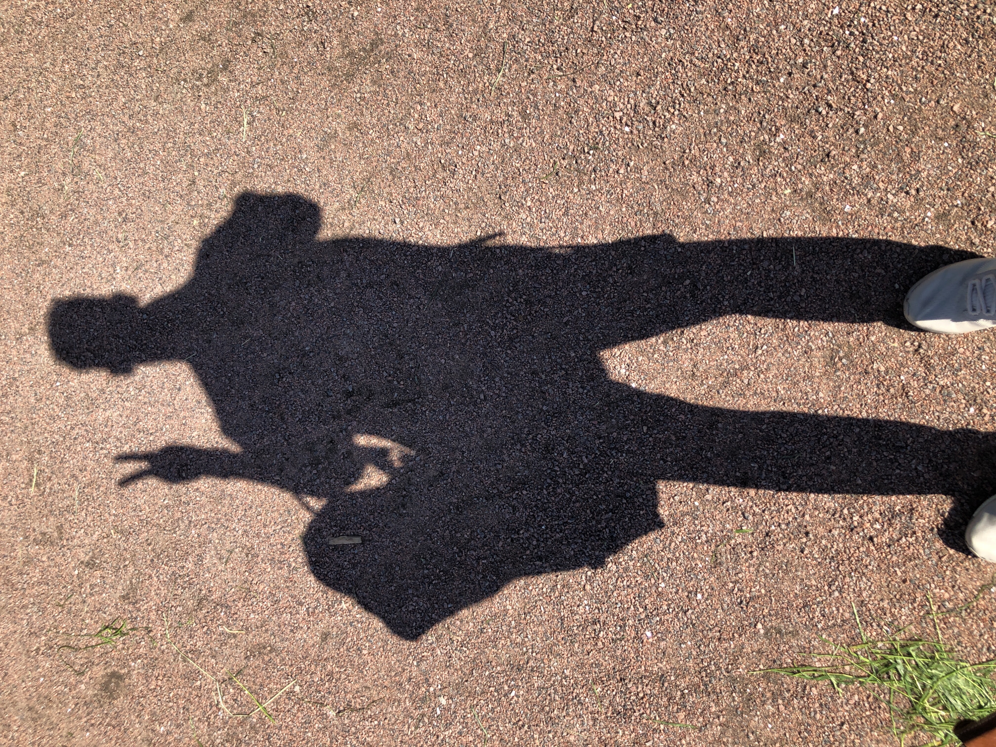 A shadow of a person is cast on a reddish-brown gravel surface. The shadow depicts a person with a backpack slung over their shoulder, holding up two fingers in a peace sign. The person's feet, clad in light-colored sneakers, are visible at the bottom of the frame. Blades of green grass are scattered across the gravel. The scene appears to be an outdoor pathway in Saint Petersburg, Russia, likely a park or public area.