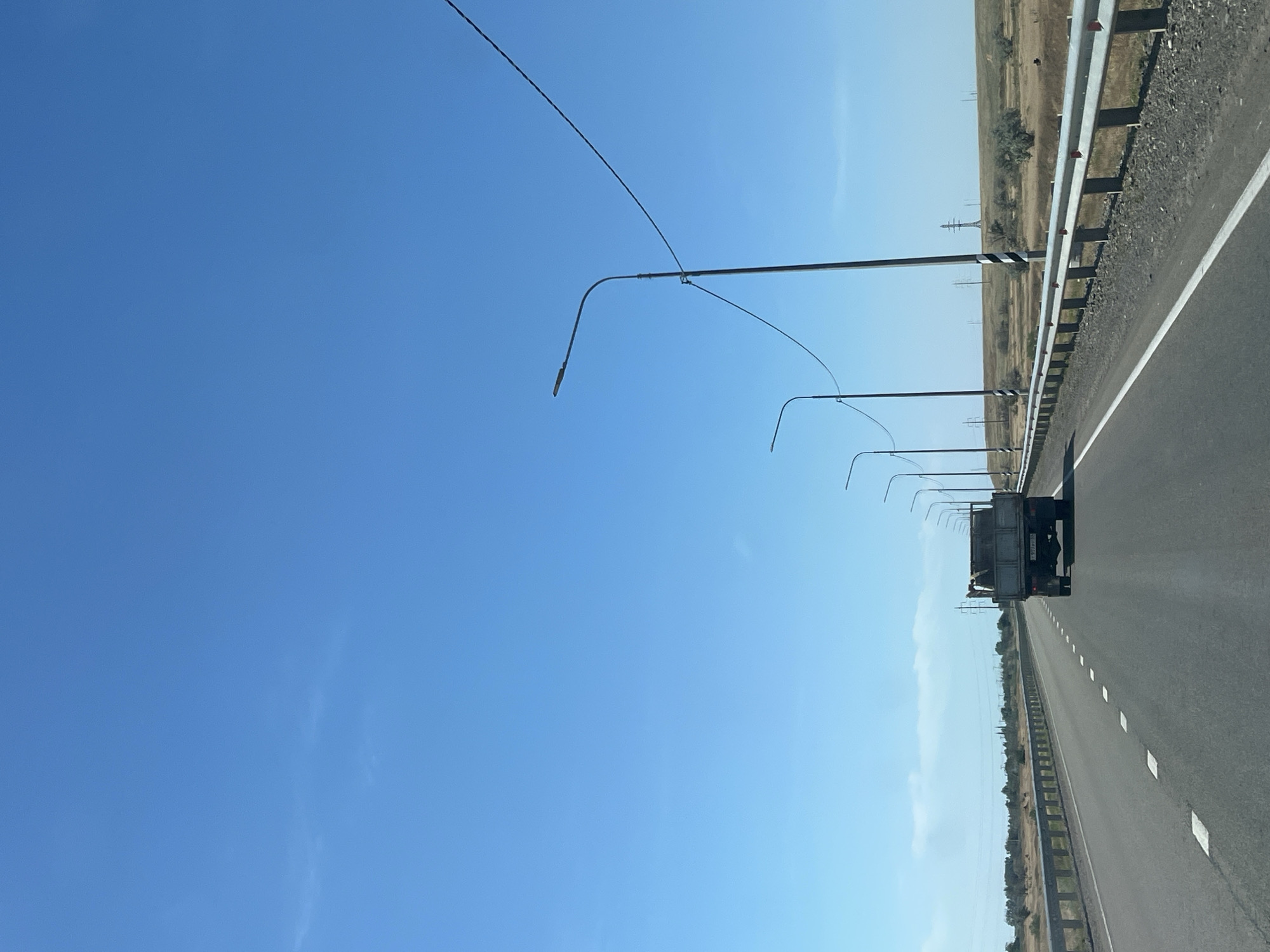 A bright, sunny day with a clear blue sky over a rural highway in Bakhtemir, Russia. A grey dump truck is driving away from the viewer down the empty road. The truck is laden with dirt or rubble. On the right side of the road, a metal guardrail runs parallel to the highway, punctuated by reflective markers. Several tall streetlights with curved arms are spaced along the guardrail, with wires connecting them. The landscape is dry and barren, with sparse, dry vegetation and a few scattered trees. In the distance, a power pylon is visible on a gentle hill. The road has white dashed lines marking the lanes. The overall impression is one of a remote, open landscape on a clear day.