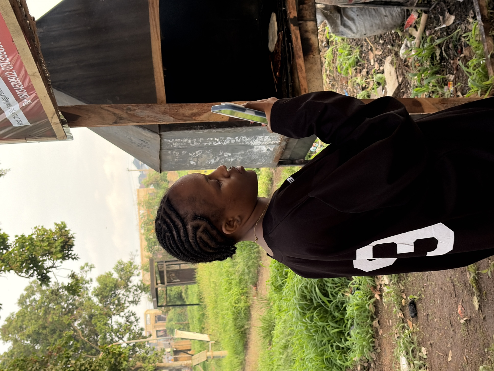 A young Black woman with braided hair is standing outdoors, looking at her smartphone. She is wearing a black long-sleeved shirt with a large white number "3" on the back. She is positioned to the left of a rustic, wooden structure that appears to be an outdoor cooking area or stall. Inside this structure, on a grate, are some cooked items, possibly flatbreads. The background shows lush green grass and some trees, suggesting a rural or semi-rural setting. In the upper right corner of the image, a sign with text and a red border is visible, attached to the wooden structure. The overall scene appears to be in Rafin Par, Nigeria, characterized by its outdoor, potentially informal commercial activity and the natural environment.