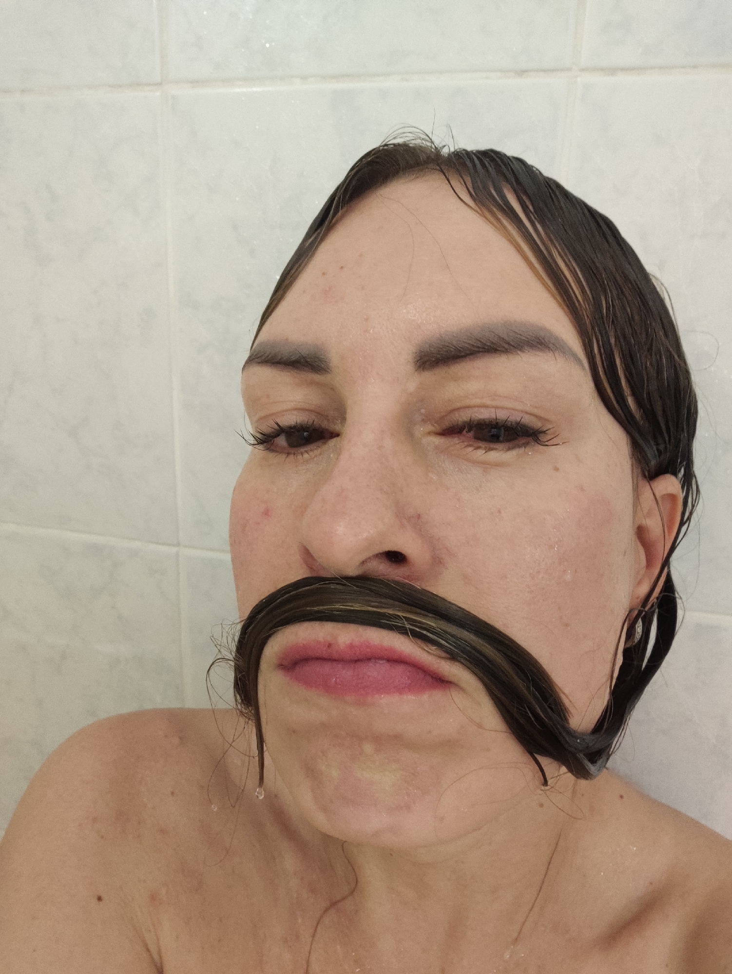 A close-up, selfie-style shot features a person in a shower, against a white tiled wall. The person's wet, dark hair is slicked back and falls over their face, forming a makeshift mustache. Their eyes are mostly closed, and their lips are slightly parted. Water droplets are visible on their skin and hair. The background is a standard tiled shower wall. There is no discernible location in the city of Almaty, Kazakhstan.