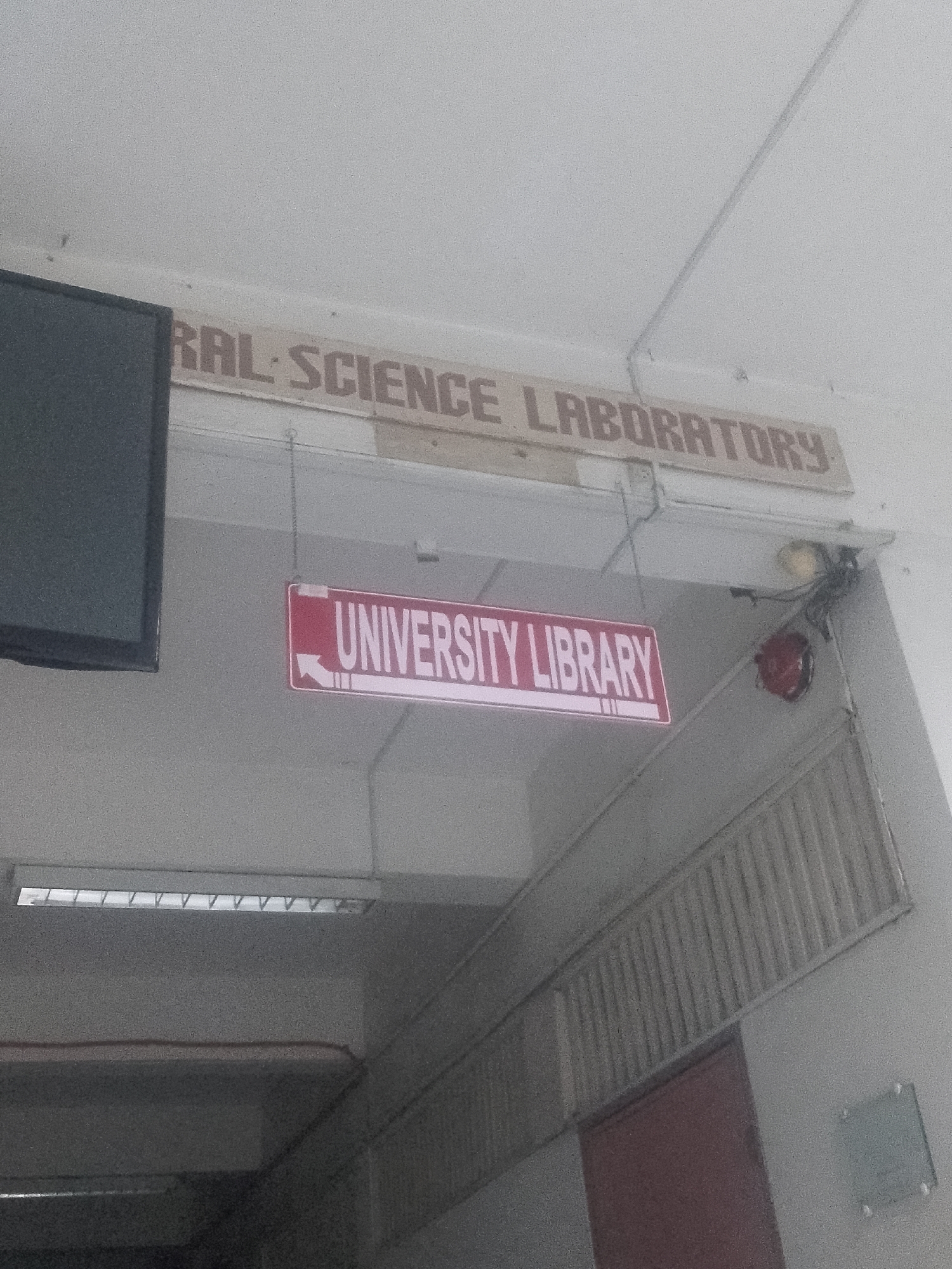The image depicts an interior corridor of a building in Davao City, Philippines. Above the doorway is a sign reading "RAL SCIENCE LABORATORY." A red and white sign hanging below indicates "UNIVERSITY LIBRARY" with an arrow pointing left. A fluorescent light fixture is visible on the ceiling. A radiator and door are visible on the right wall. An electronic display screen is visible on the left side of the image.
