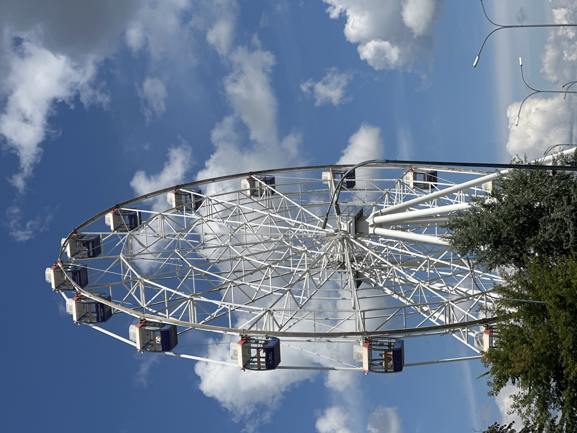 A white Ferris wheel stands tall against a bright blue sky filled with scattered white clouds. Several blue and white gondolas are attached to the wheel, with some visible passengers inside. The Ferris wheel is framed by lush green trees in the foreground, and streetlights are visible in the background. The scene appears to be an outdoor amusement park or recreational area.