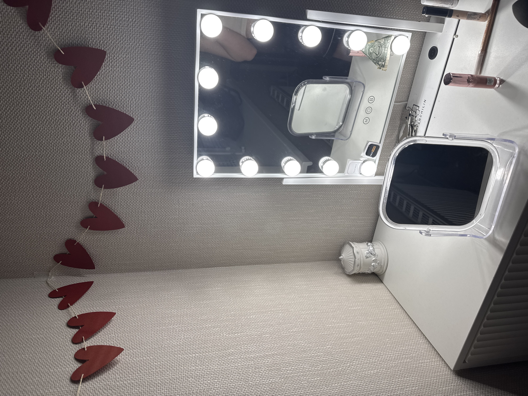 The image shows a close-up of a vanity area. On the left, a string of red paper hearts hangs against a light-colored, textured wall. Centered, a makeup mirror with lights borders its edge, reflecting parts of the room including a human body. Adjacent to the mirror, makeup products are arranged. Further to the right stands another mirror with a plastic protective case, partially obscuring its view, next to a small decorative carousel sculpture. The scene is well-lit, and the surface of the vanity is a light gray fabric. It's not possible to discern the specific location within the city of Perm, Russia from this single image.