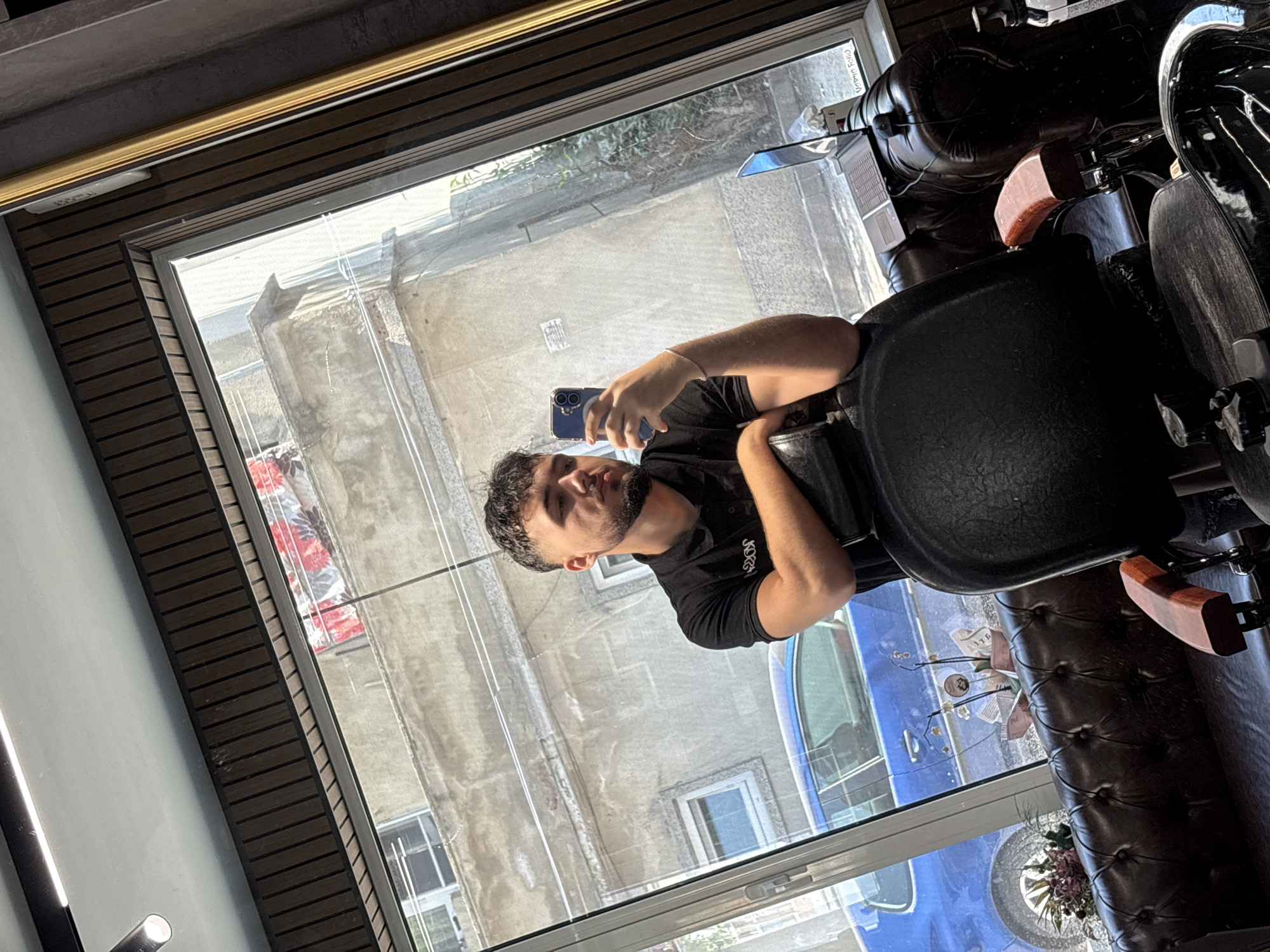 An image, rotated 90 degrees clockwise, captures a young man taking a selfie inside what appears to be a barbershop in Varna, Bulgaria. The man, with dark, curly hair and a beard, is wearing a black collared shirt, partially revealing the word "JOKER" on it. He is seated in a dark, possibly leather, chair and holds a dark blue smartphone with a distinctive case up to capture his reflection in a large window. A silver-colored bracelet adorns his left wrist.
The interior setting features a prominent window frame with dark brown slats and a golden decorative trim. To the right, a dark brown tufted leather bench or chair holds a small bouquet of artificial pink/purple flowers and some papers. Behind the man, a silver laptop is visible on a surface, reflecting in the window.
Through the window, under bright daylight, an urban street scene is visible. Old, weathered buildings with numerous windows line the street, and a blue car is parked outside. Green foliage and trees are reflected in the upper sections of the window.
Visible text provides further details: The inverted reflection of "BARBER SHOP" can be seen on the interior window frame, confirming the establishment. Papers on the leather bench display Bulgarian text, including "МАГАЗИН" (Shop), "ЛИМИТ" (Limit), and "ПАРФЮМЕРИЯ" (Perfumery), reinforcing the Bulgarian context. The bright lighting suggests it is daytime.
